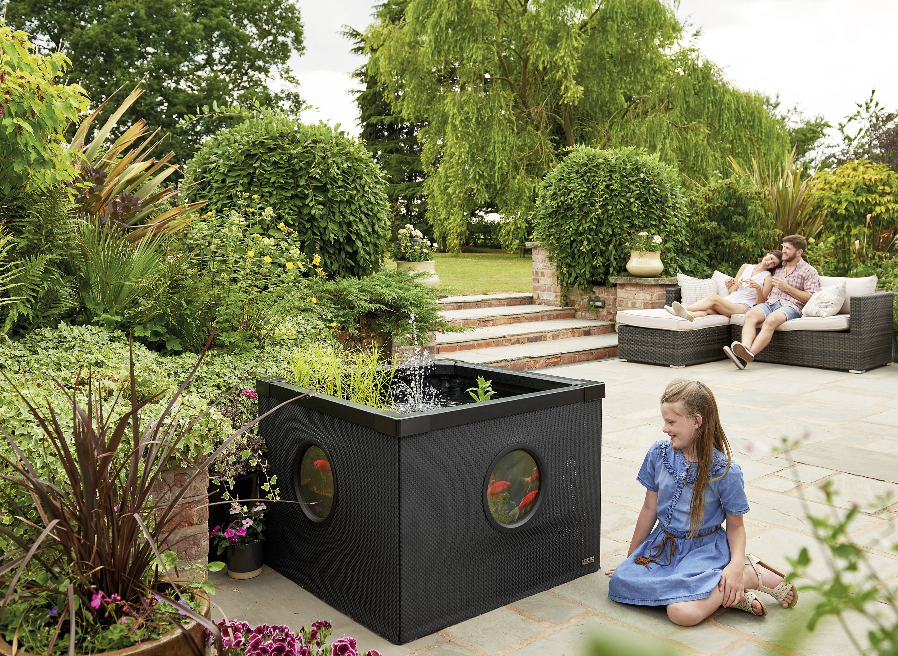 A girl sits in the garden in front of a small rectangular garden pond with fish. A couple relaxes on garden furniture in the background.