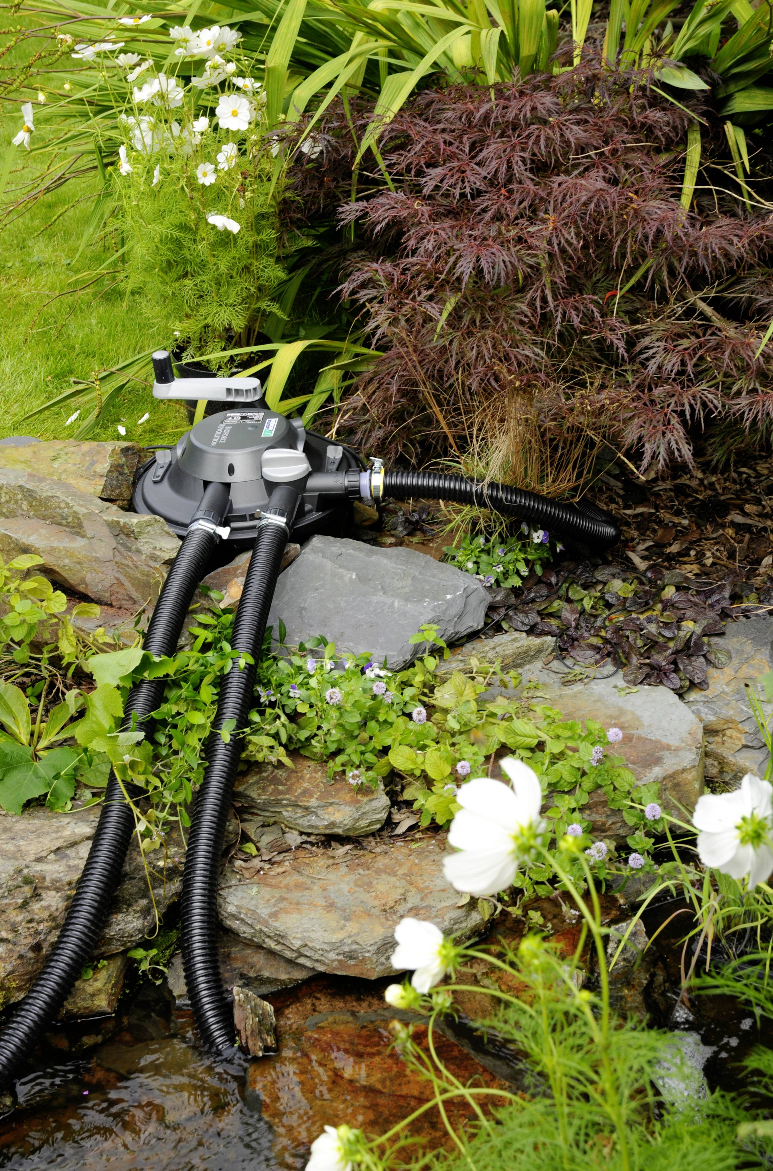 Pond pump with black hoses and control elements, positioned on a rock amidst lush, green vegetation.