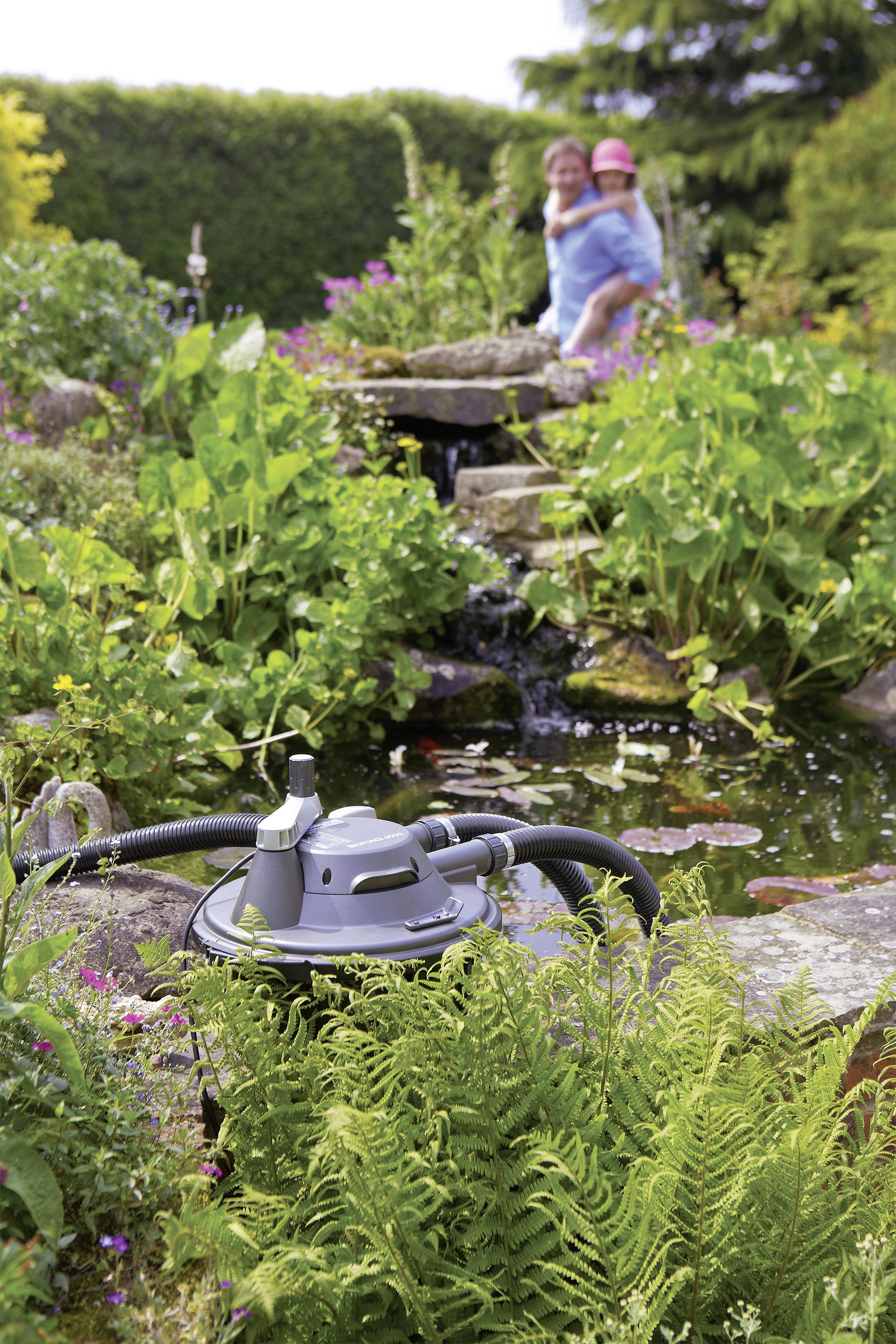 A pond pump in front of a garden pond with lush vegetation; in the background, a person carrying a child.