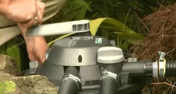 A person is adjusting a garden pond filter with a pipe wrench. The filter is circular and connected to several hoses.