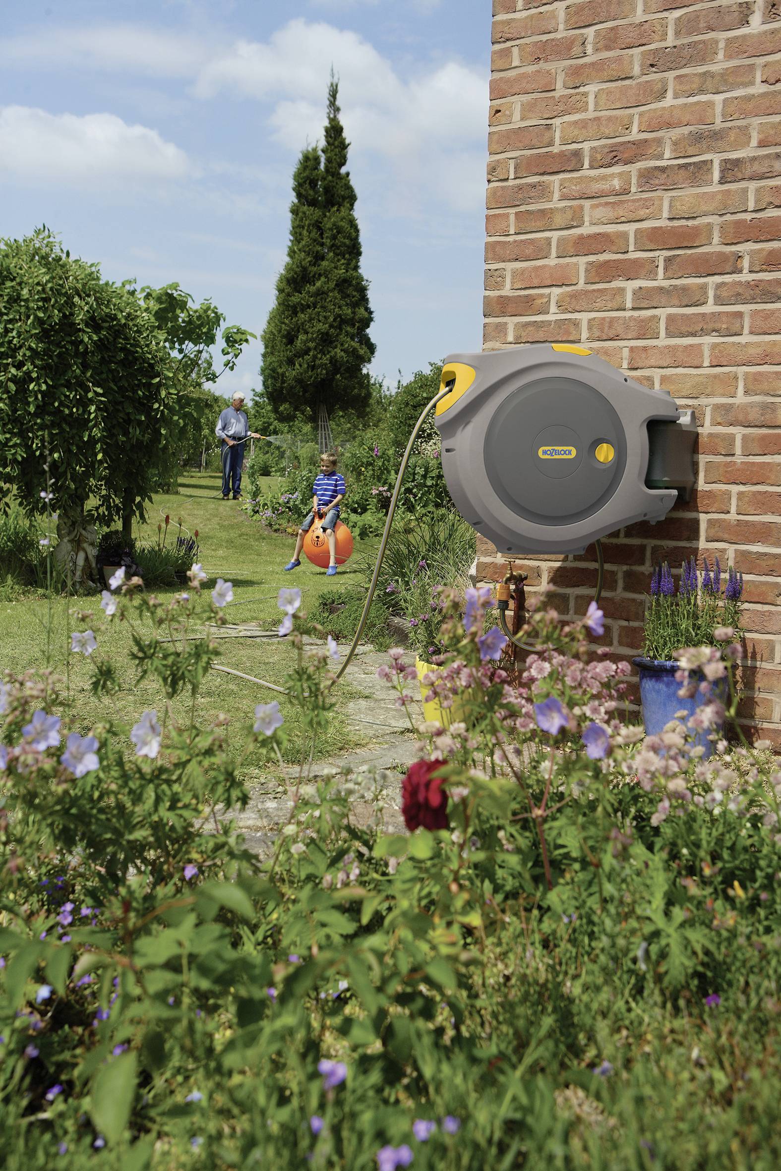A garden scene with a hose reel hanging on a brick wall, with a child on a space hopper and an adult in the background.