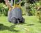 A person is pulling a grey hose trolley with yellow details across a green lawn in a garden with flower beds.