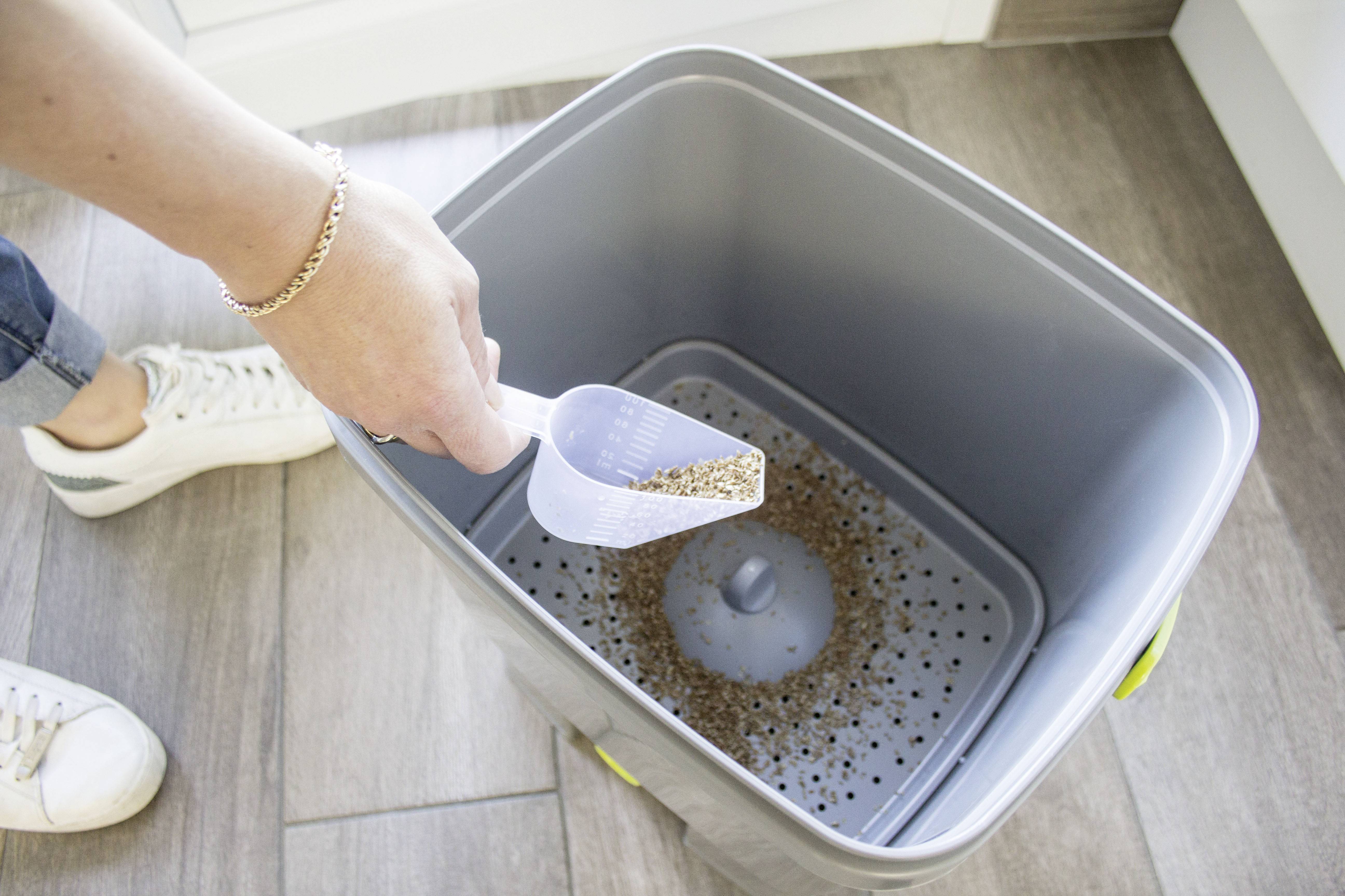 'A person is scattering pellets into a grey compost bin with a perforated base, standing on a wooden floor. A shoe is visible on the left.'