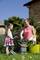 Mother and daughter smile while working in the garden. They collect pruned plants in buckets. A stone wall and a house are in the background.