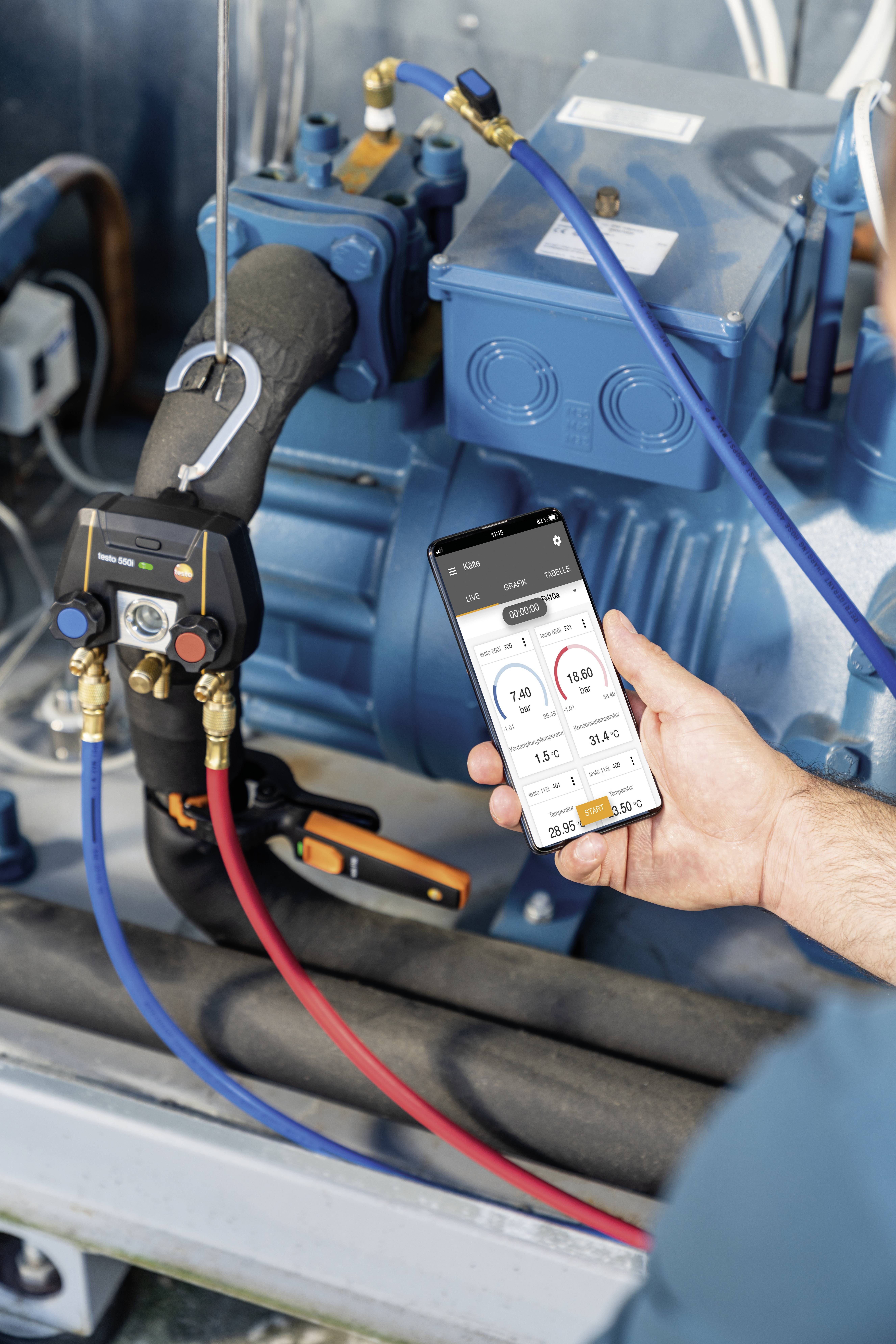 A person is holding a smartphone displaying temperature and pressure readings in front of an industrial machine with blue and red pipes.