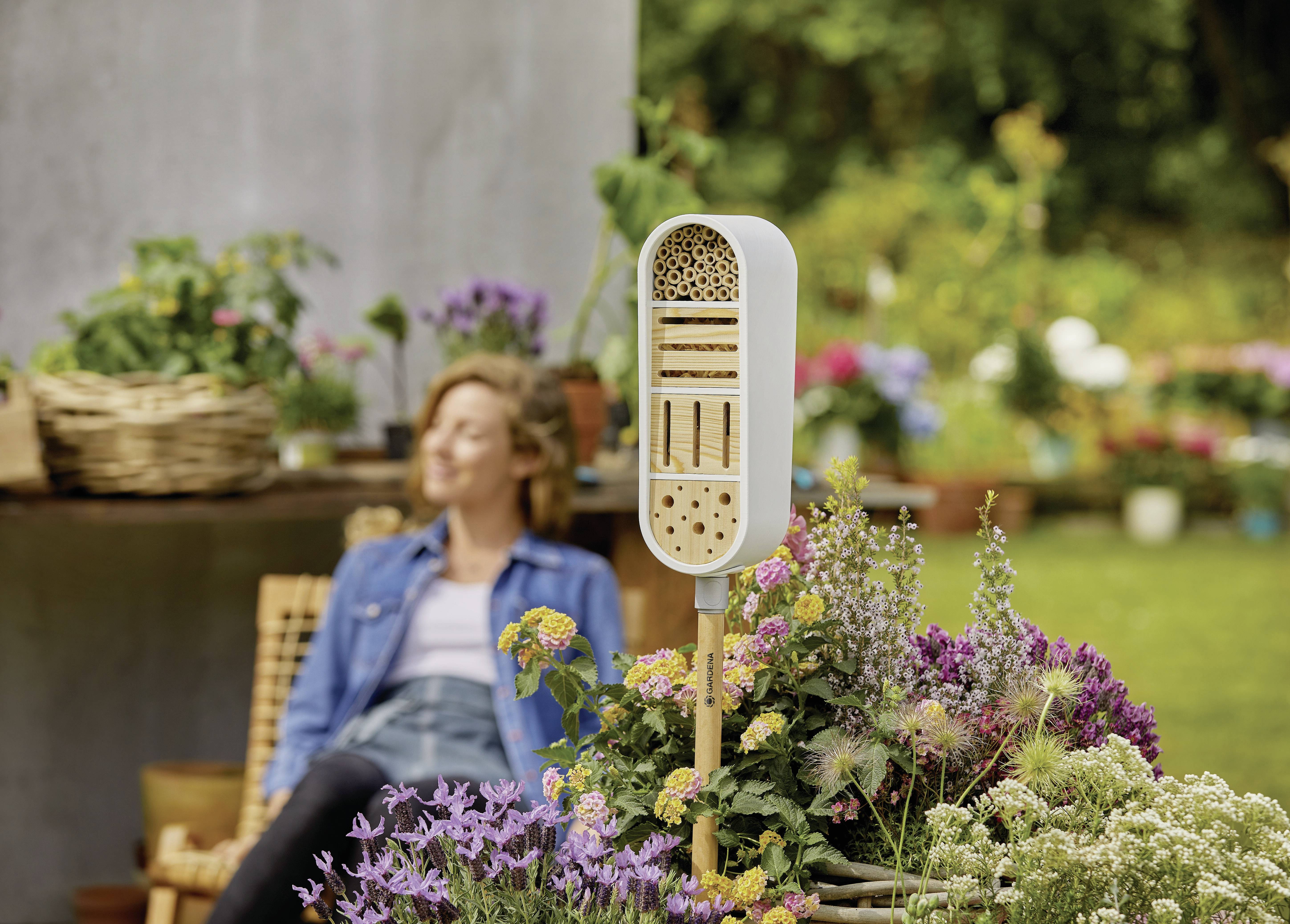 A woman sits relaxed on a chair in the garden. In the foreground, there is an insect hotel and flowering plants.