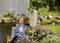 A woman sits relaxed on a chair in the garden. In the foreground, there is an insect hotel and flowering plants.