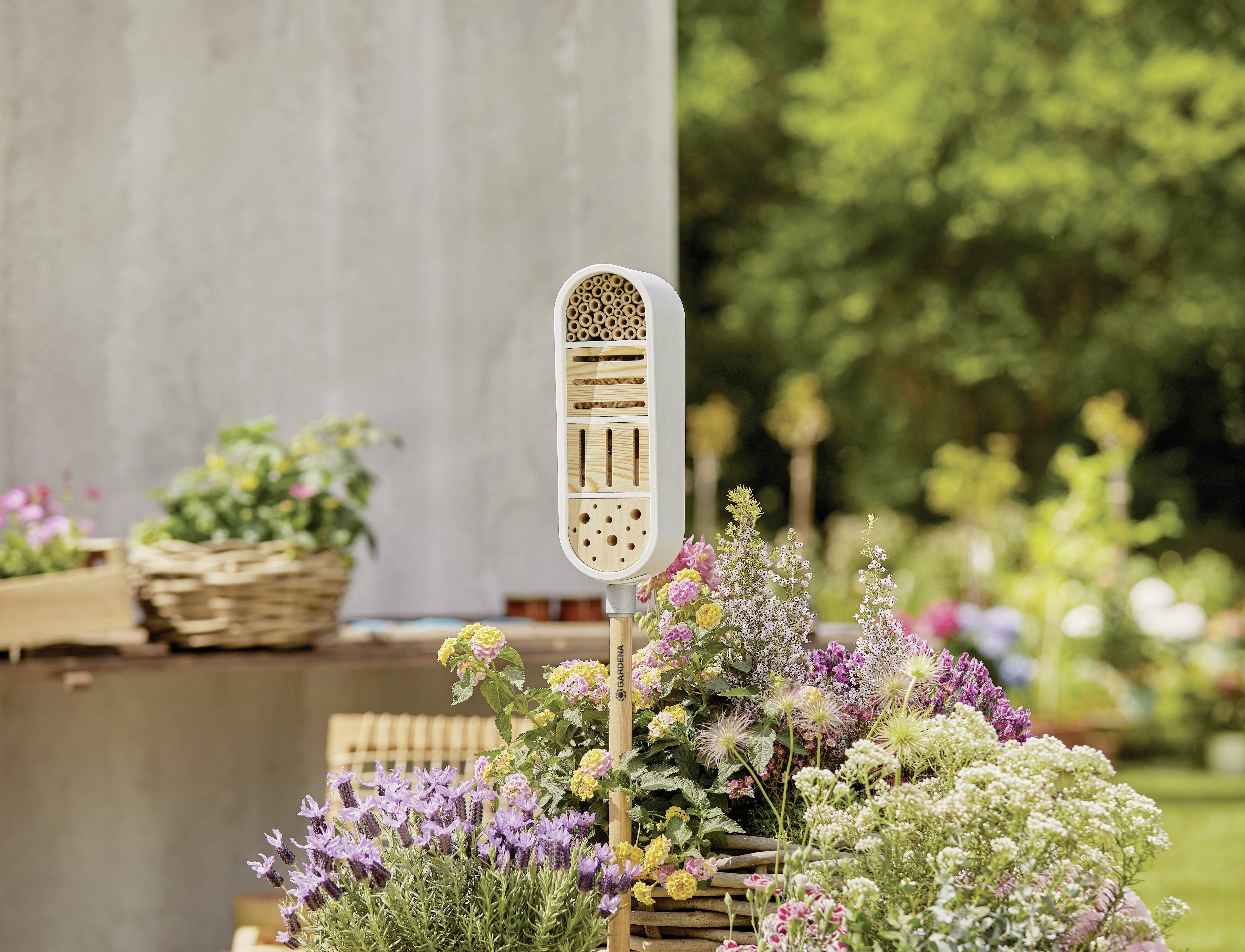 A decorative insect hotel stands in a blooming garden, surrounded by colourful flowers and plants, serving as a nesting aid.