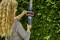 A person is trimming a green hedge with an electric hedge trimmer. They are holding the device securely with both hands.