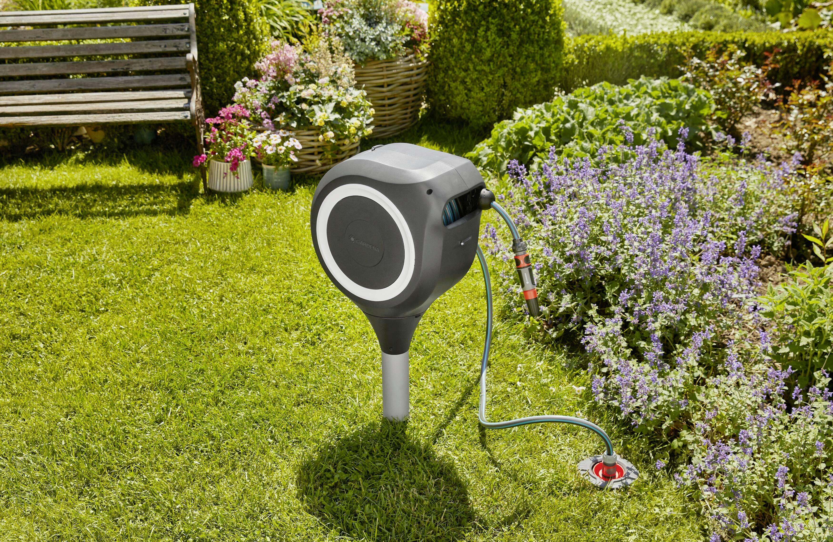 A coiled garden hose box sits in a green area beside flowering plants. A bench and colourful flowers can be seen in the background.