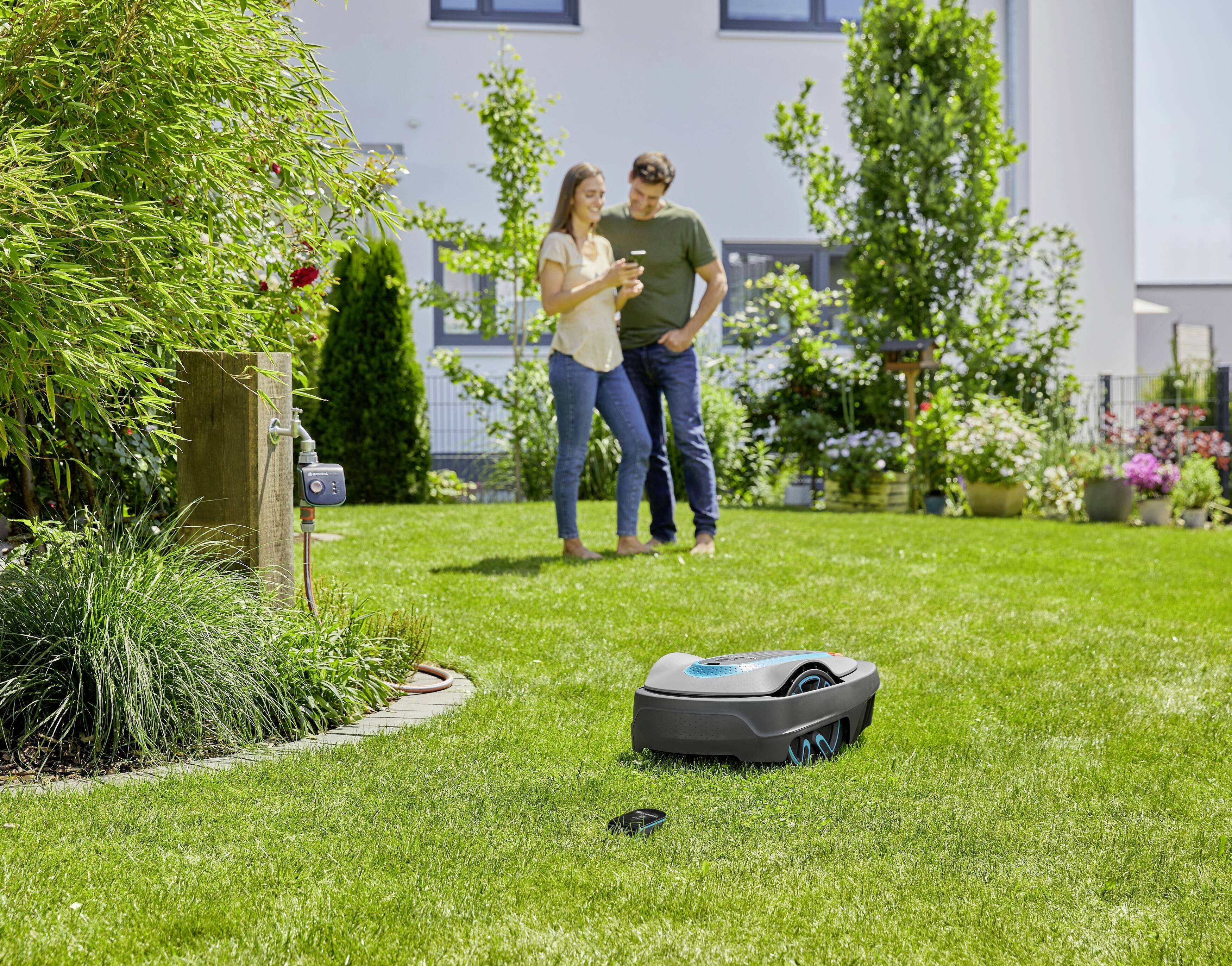 A couple stands smiling in the garden, looking at a tablet while a robotic lawnmower cuts the grass. Flowering plants bloom in the background.