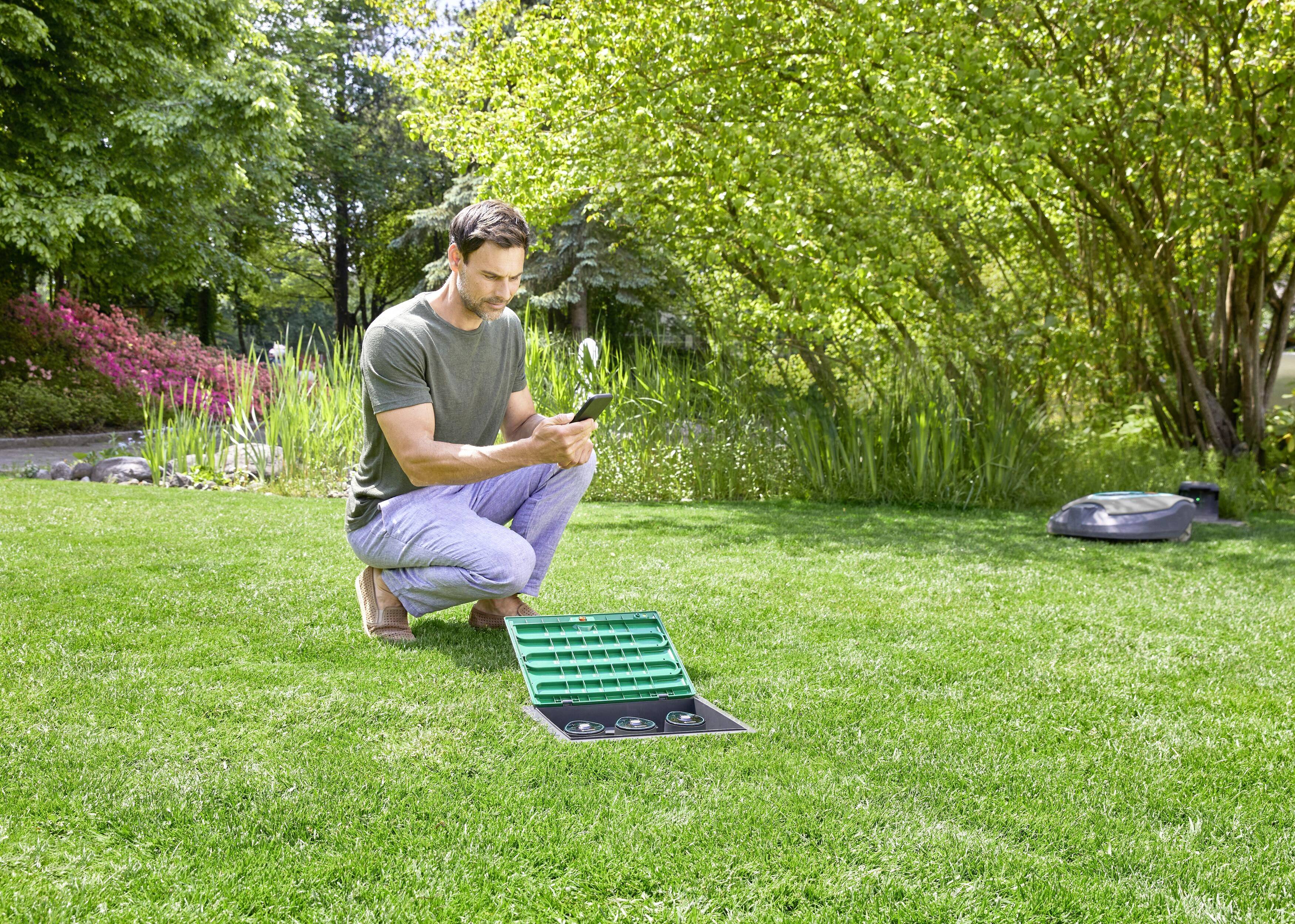 A man is kneeling on a grassy area and controlling an irrigation system via his smartphone. Trees and bushes are visible in the background.