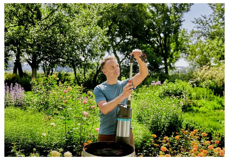 Man outdoors in a sunny garden adjusting a stainless-steel device or pump beside a flower bed.