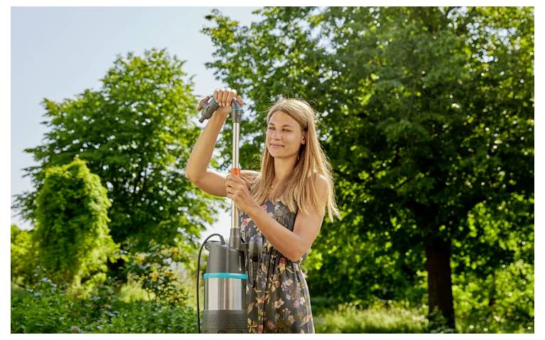 Woman using a hand water pump or garden sprayer outdoors in a sunny green garden.