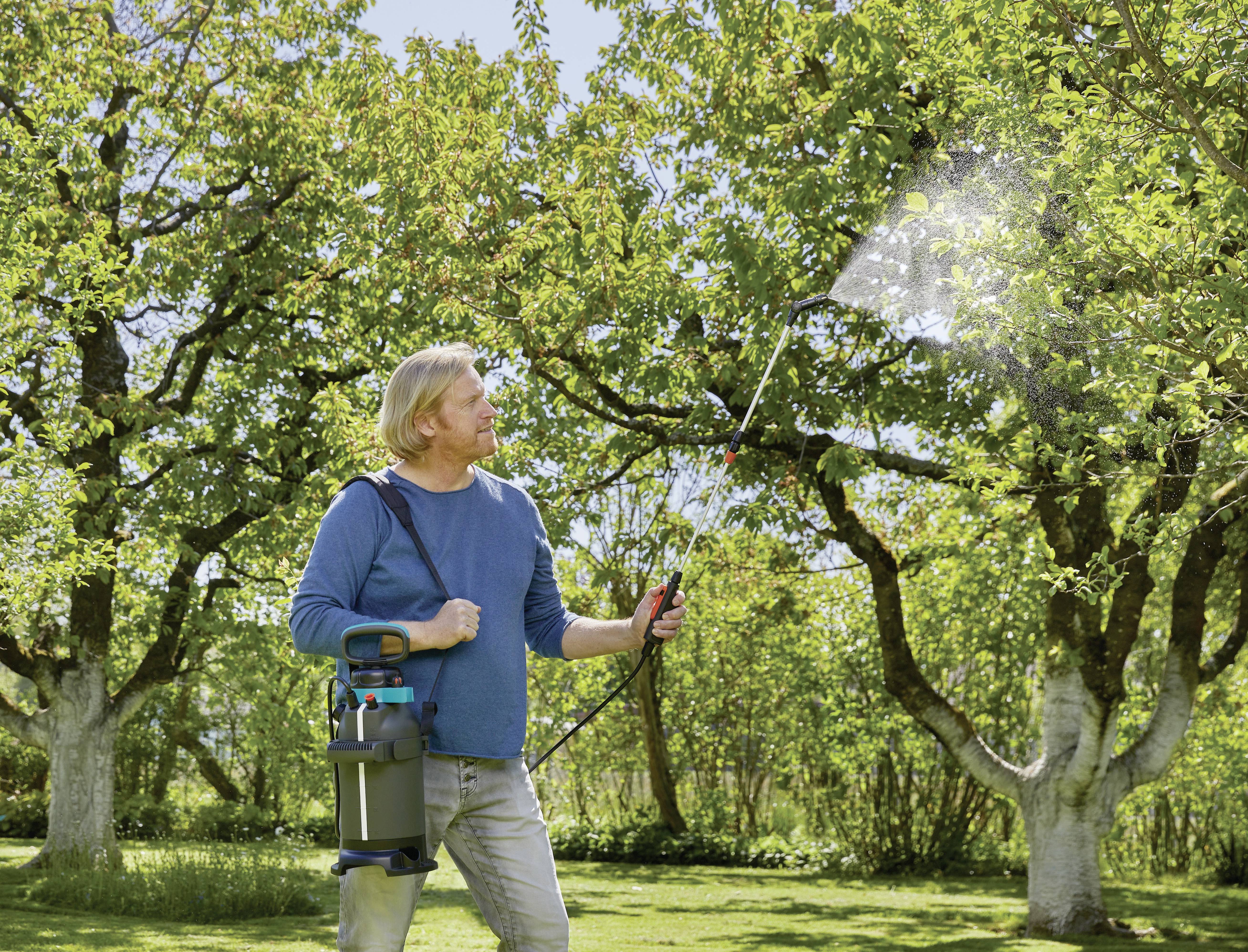 A person is spraying liquid onto a tree with a portable spraying device in a green garden.