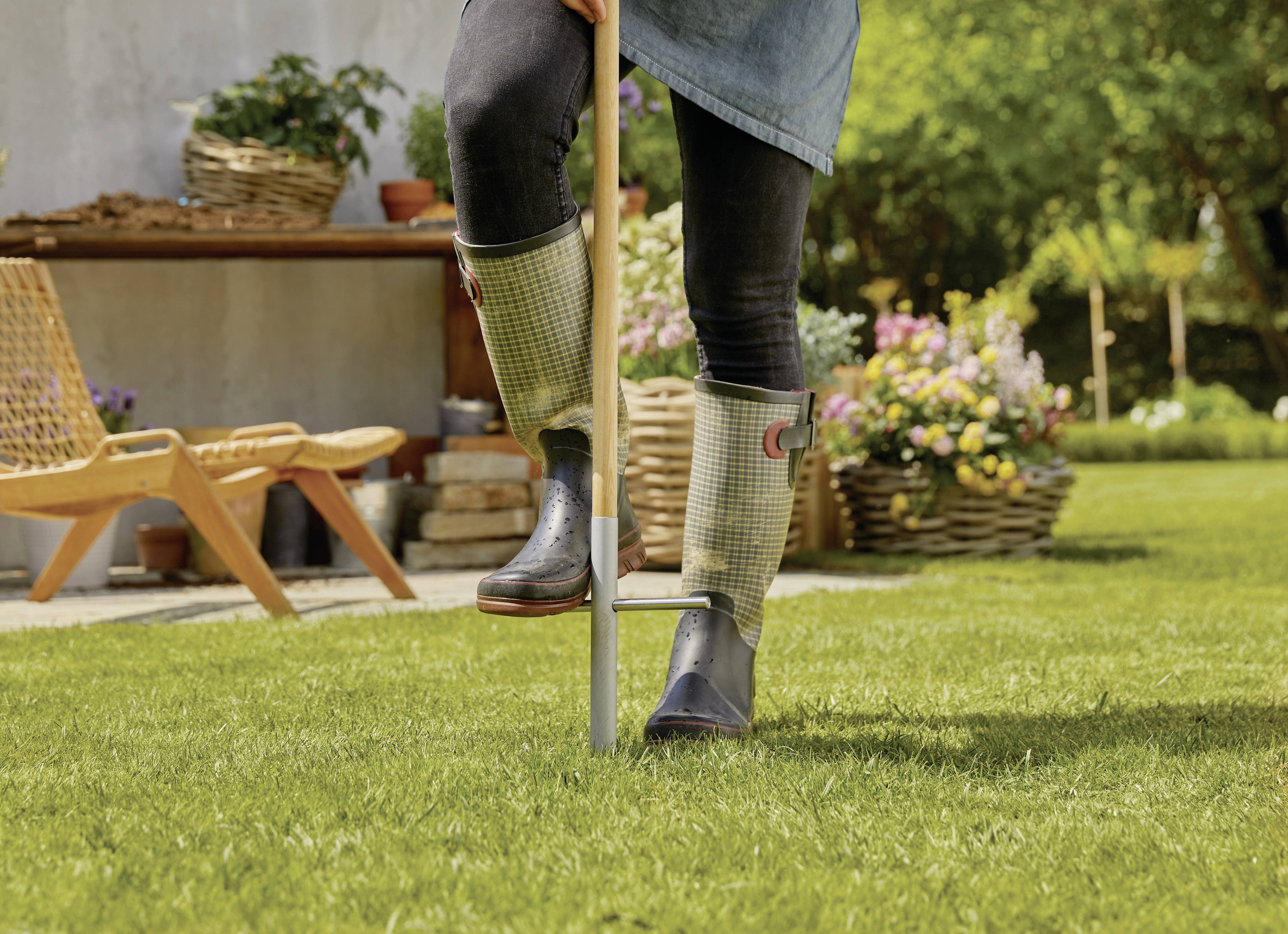 A person wearing wellington boots is using a garden tool on a lawn. Flowers and a table are visible in the background.