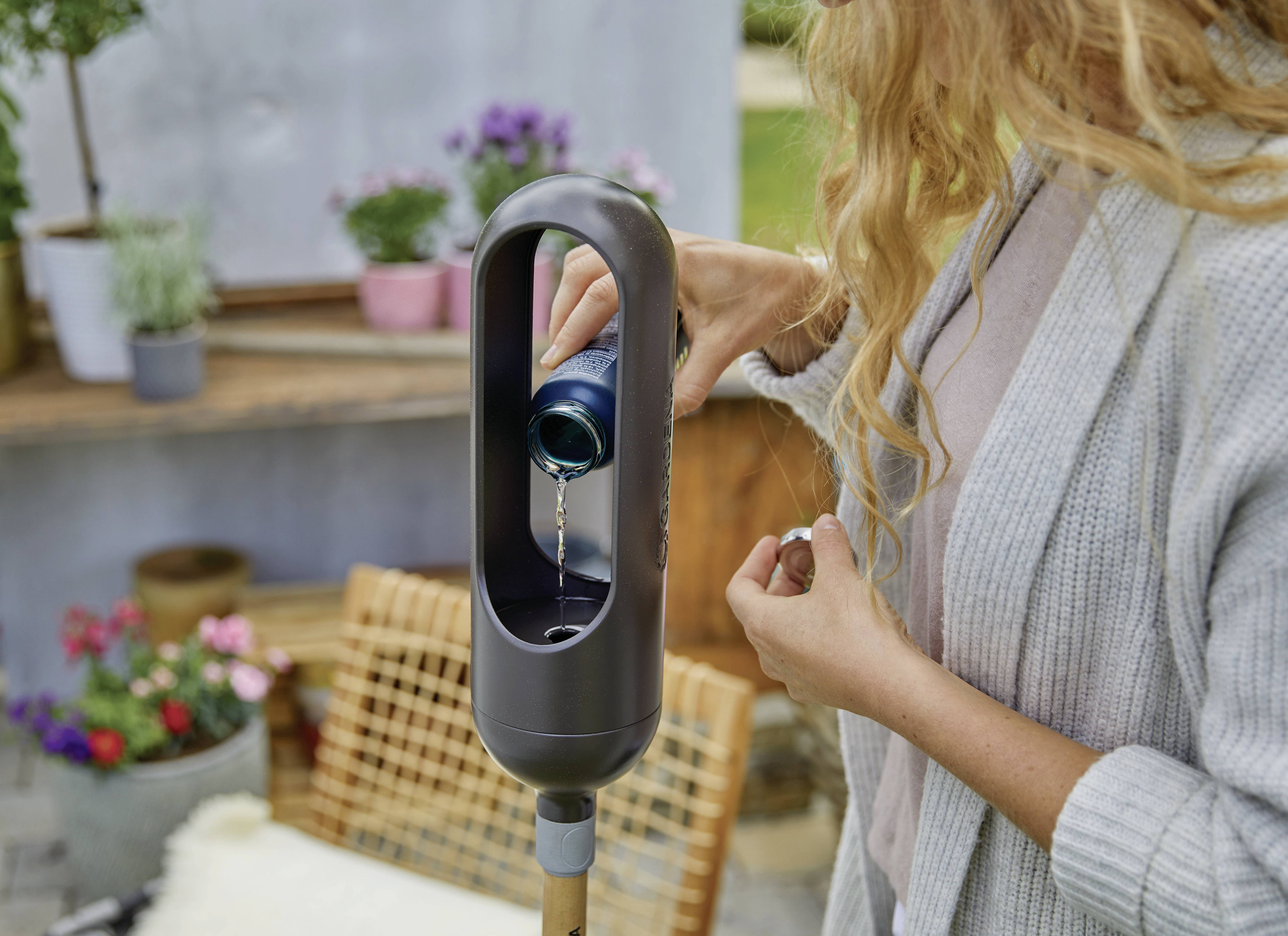 A person is pouring a drink can into a fan-like device outdoors. Garden plants are visible in the background.