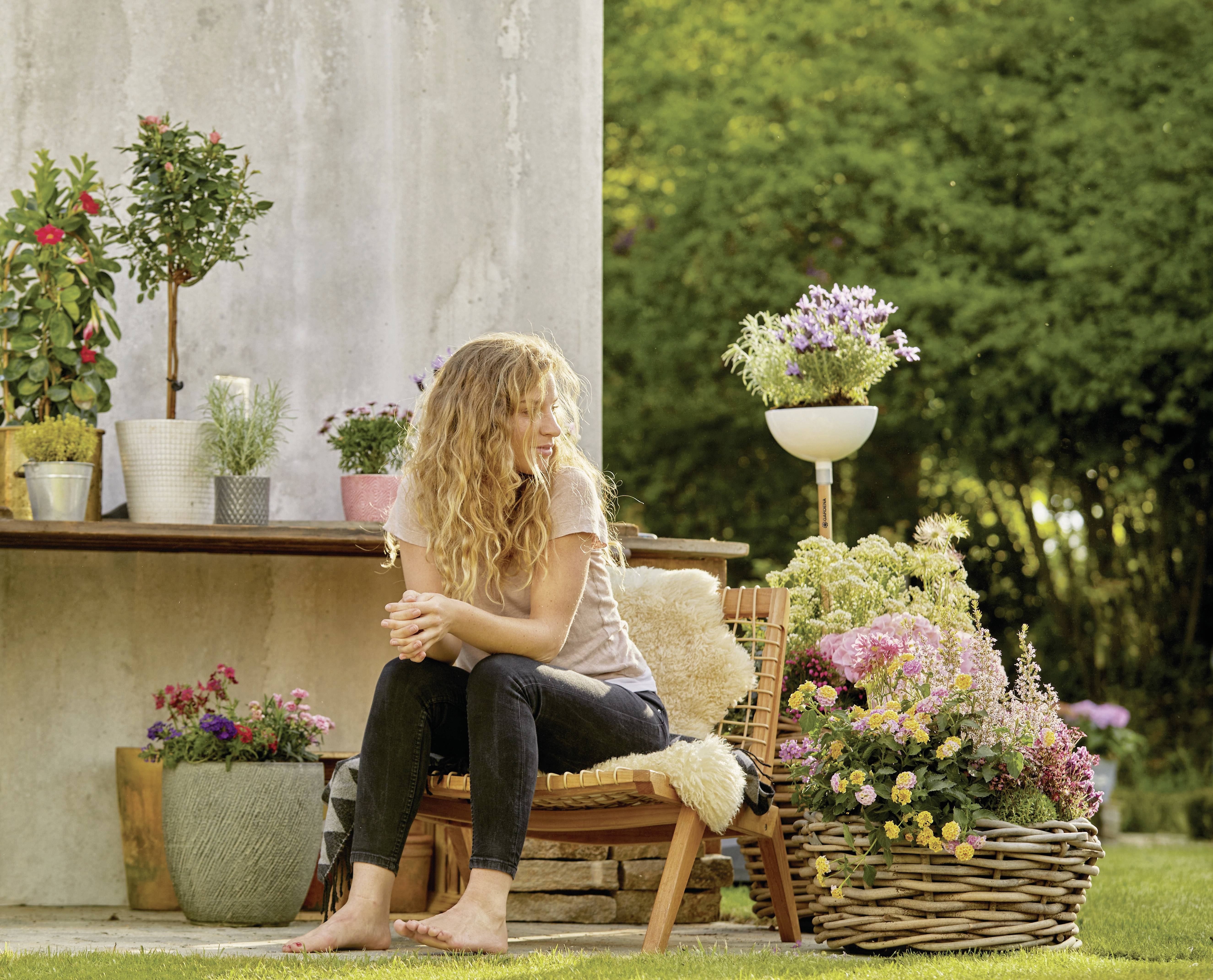 A woman with curly hair sits outside on a chair, surrounded by blooming plants and colourful flowers, in front of a garden wall.