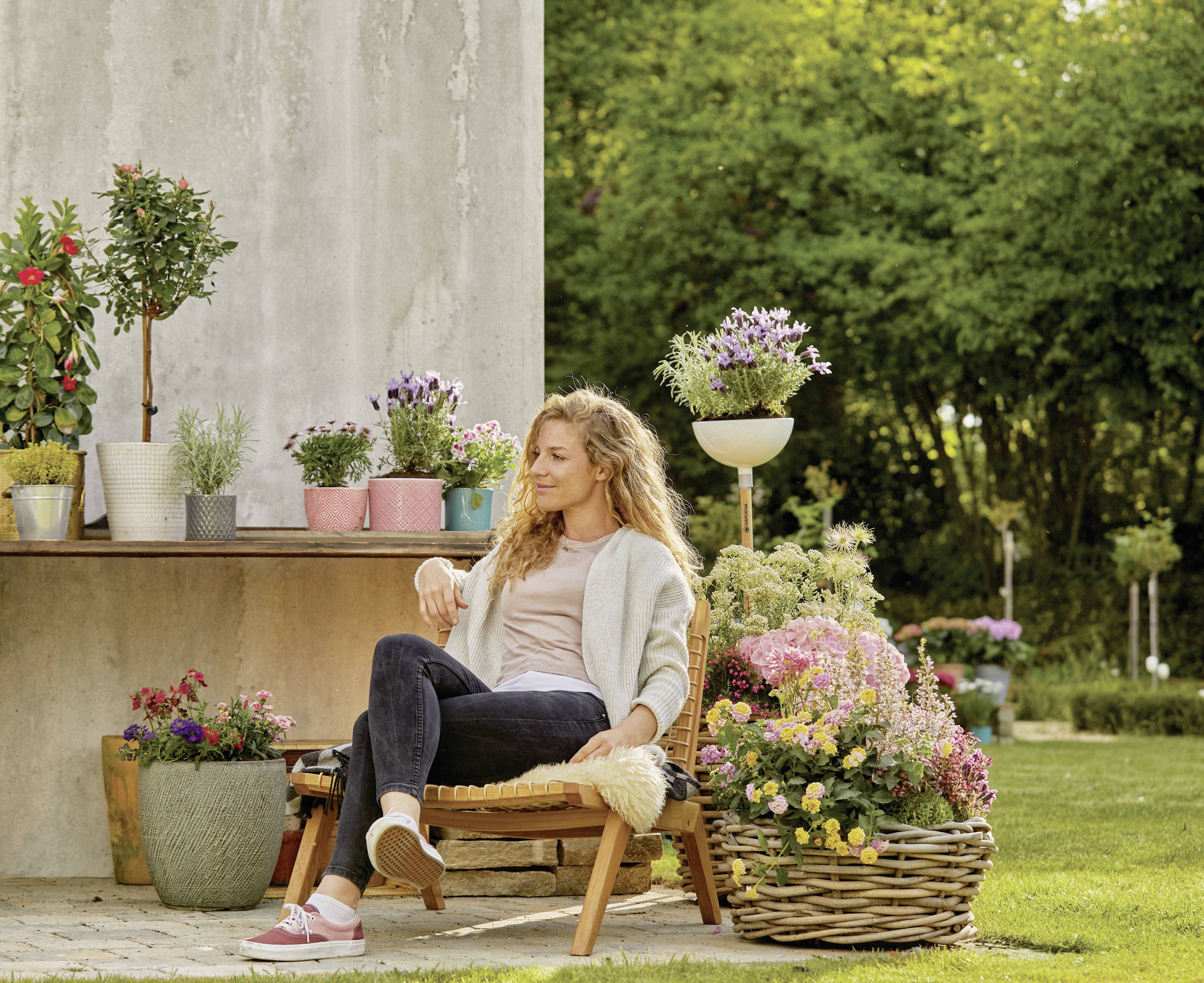 A woman sits relaxed on a chair in the garden, surrounded by blooming plants and flowers. In the background, a table with flower pots is visible.