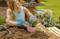 A woman wearing gardening gloves is planting flowers in a plant pot on an outdoor table, surrounded by soil and gardening tools.