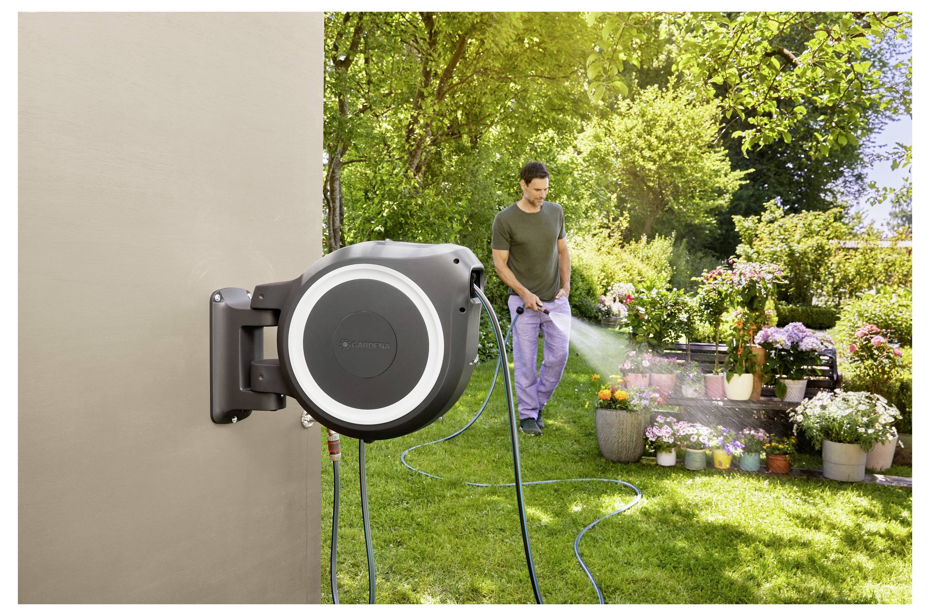 A person waters a garden with a hose connected to a wall-mounted retractable reel. They are surrounded by plants and flowers in pots.