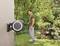 A man is standing in a garden holding a hosepipe attached to a wall mounting. Plants can be seen in the background.