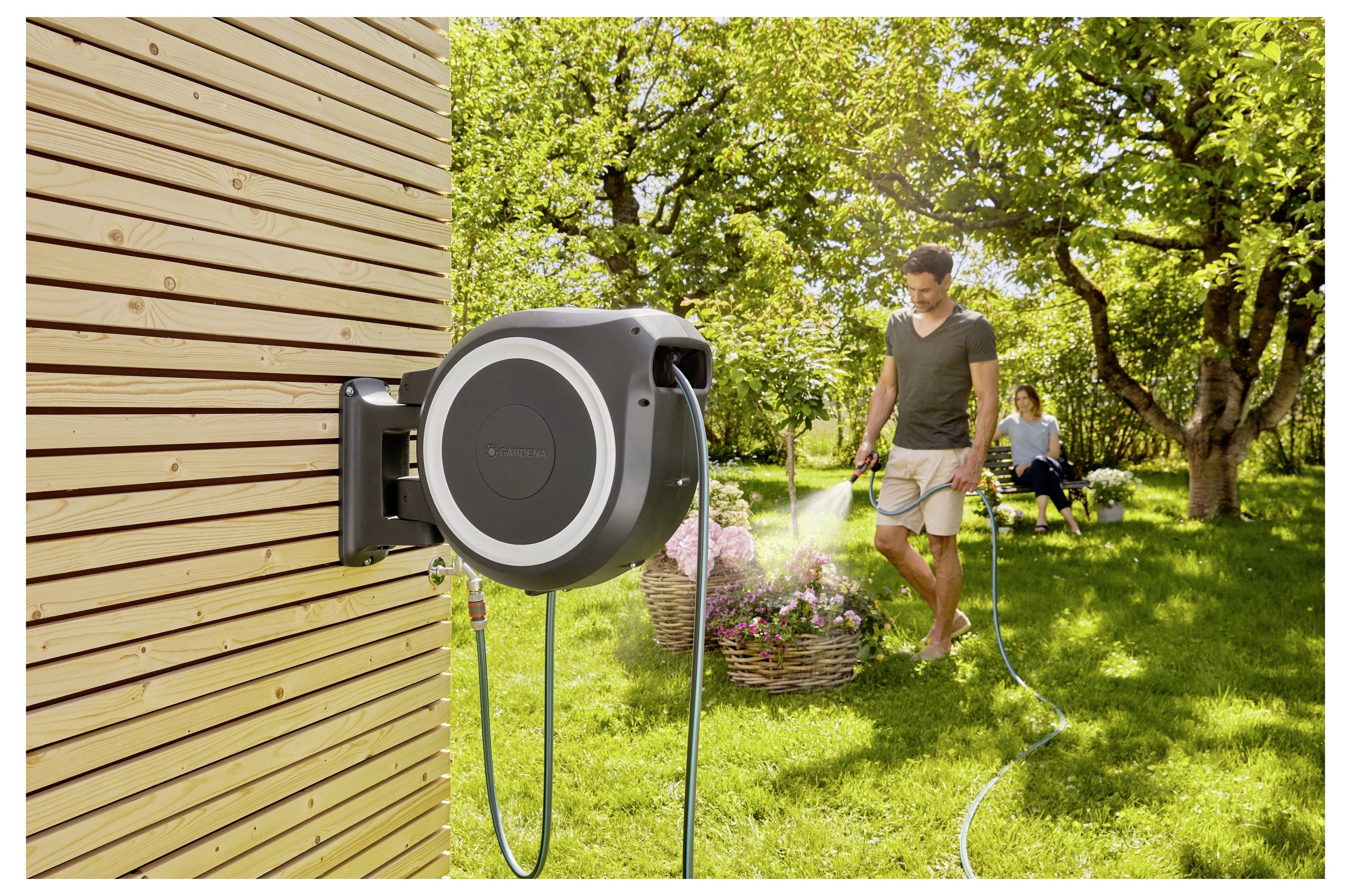 A man waters a garden using a hose reel attached to a wooden wall. A woman sits nearby among flowers, in an orchard setting.