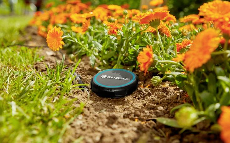 A black Gardena sensor is placed in a garden bed surrounded by orange flowers and green grass, monitoring soil conditions.