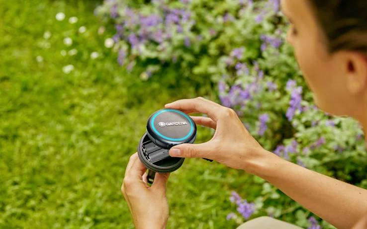 A person holds a round device labeled 'GARDENA' near a garden with blooming lavender, suggesting garden care or maintenance use.