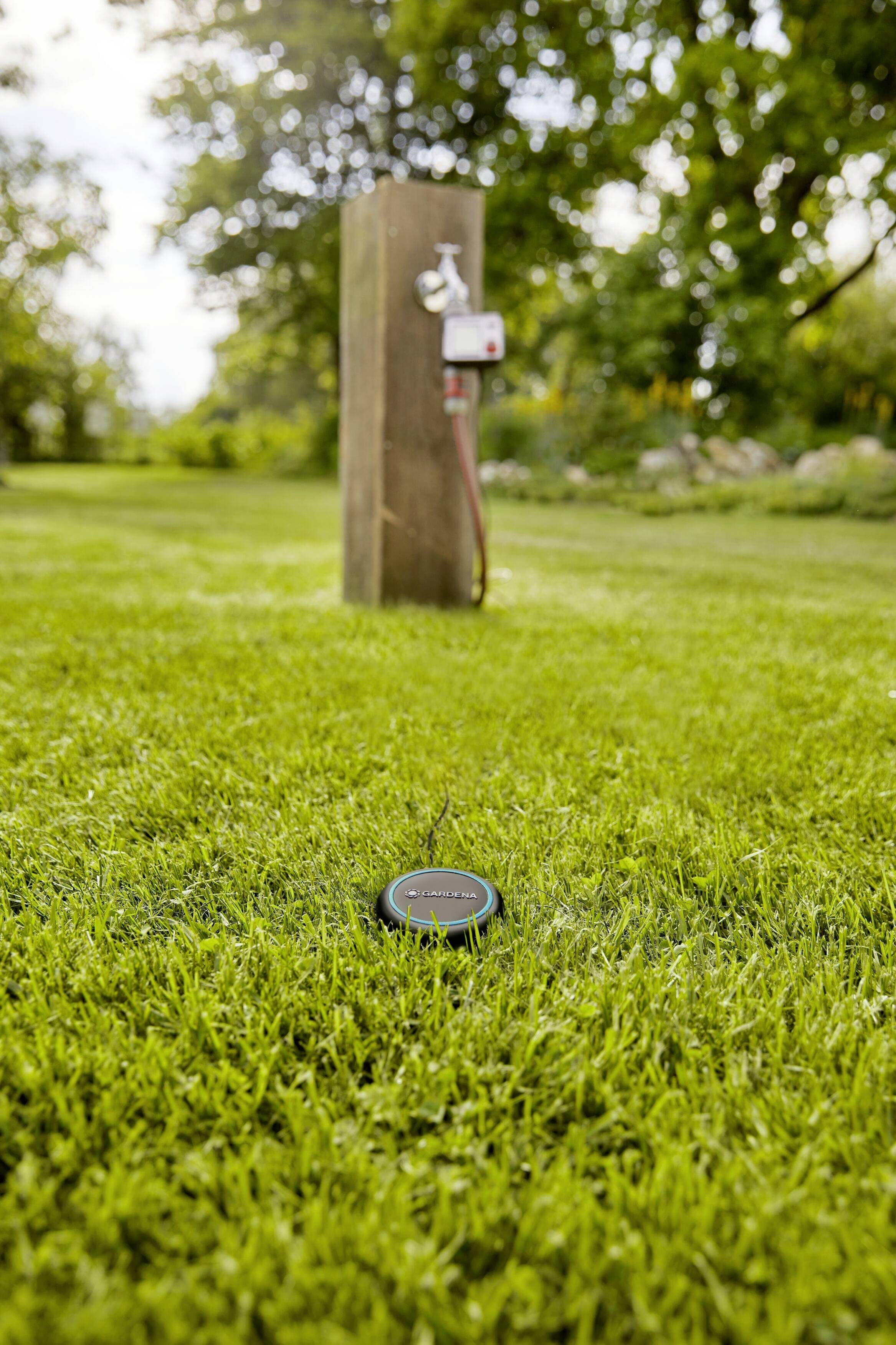 A sensor lies in the grass in front of an irrigation system on a meadow. In the background, a wooden post with a tap can be seen.