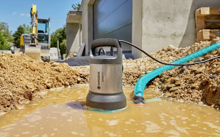 A submersible pump is draining water from a puddle on a construction site. An excavator can be seen blurred in the background.