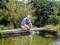 A person is kneeling by the pond's edge in a garden and using a water pump to fill the pond. Trees and bushes are visible in the background.
