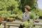 A woman sits at a wooden table in the garden and waters plants in small pots. Green trees and bushes are visible in the background.