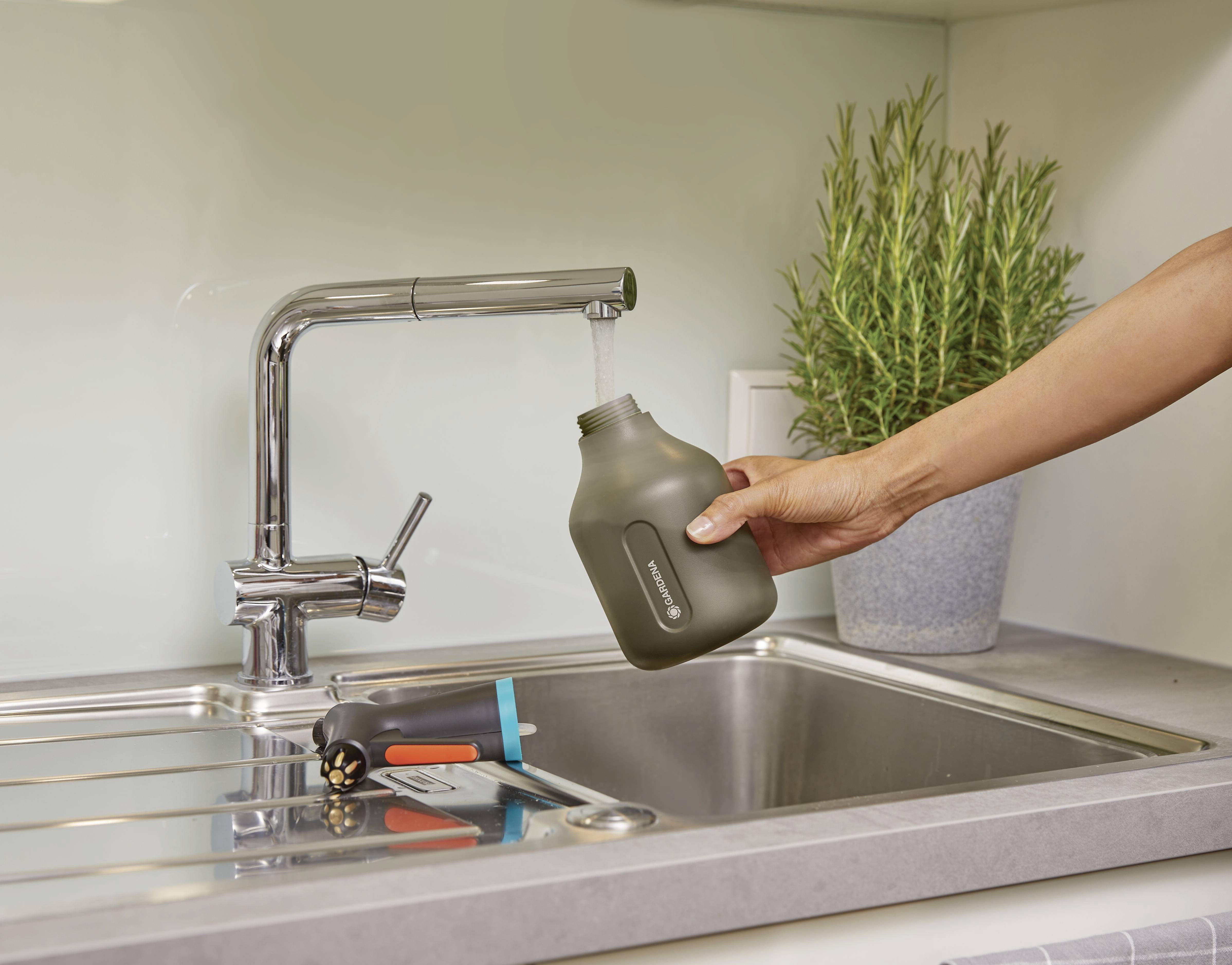 A hand holds a grey water bottle under a running tap in a kitchen. Beside it lie a black watering can and a green plant.