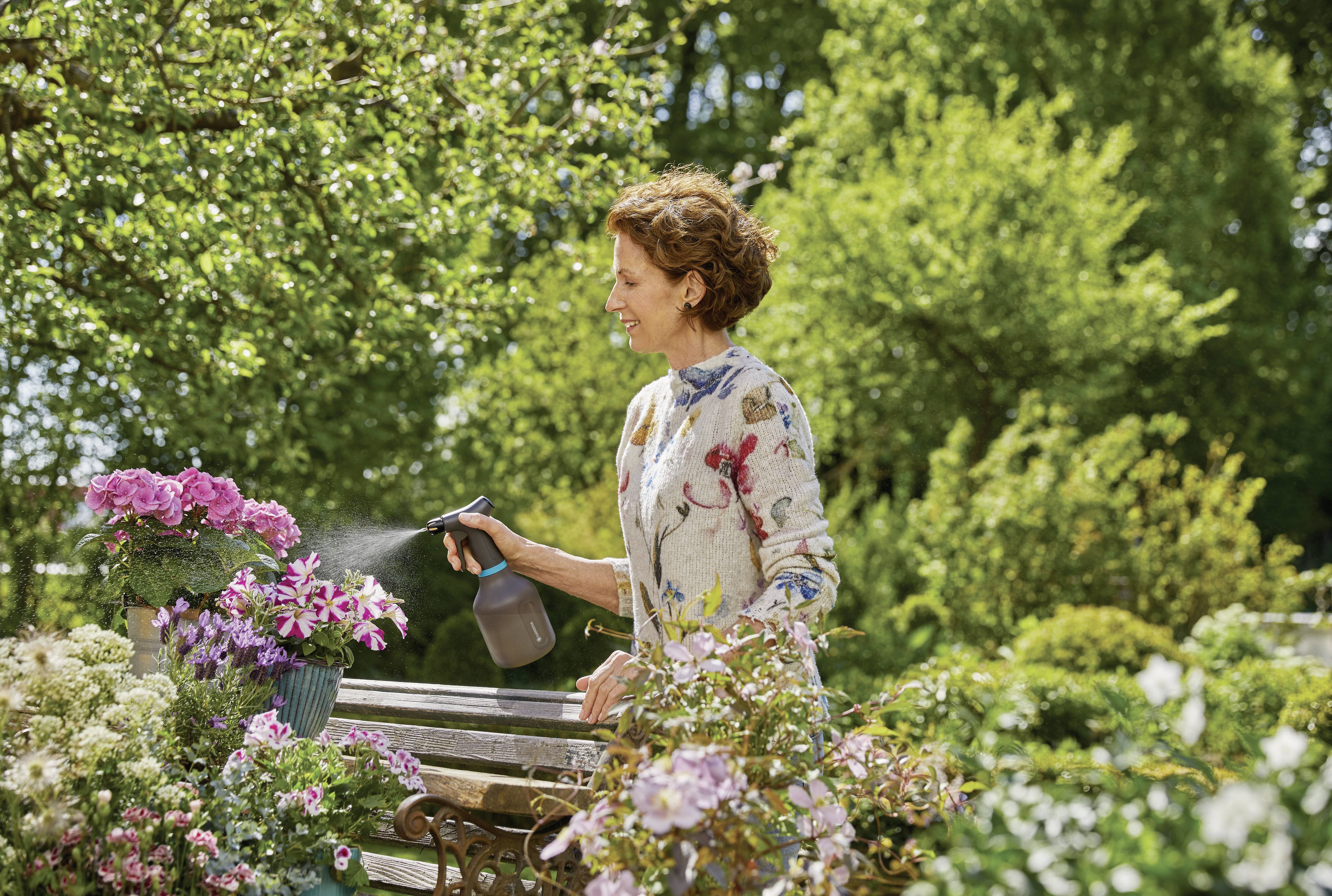 A woman in the garden is spraying flowers with water. She is wearing a floral blouse. Around her are green trees and blooming plants.