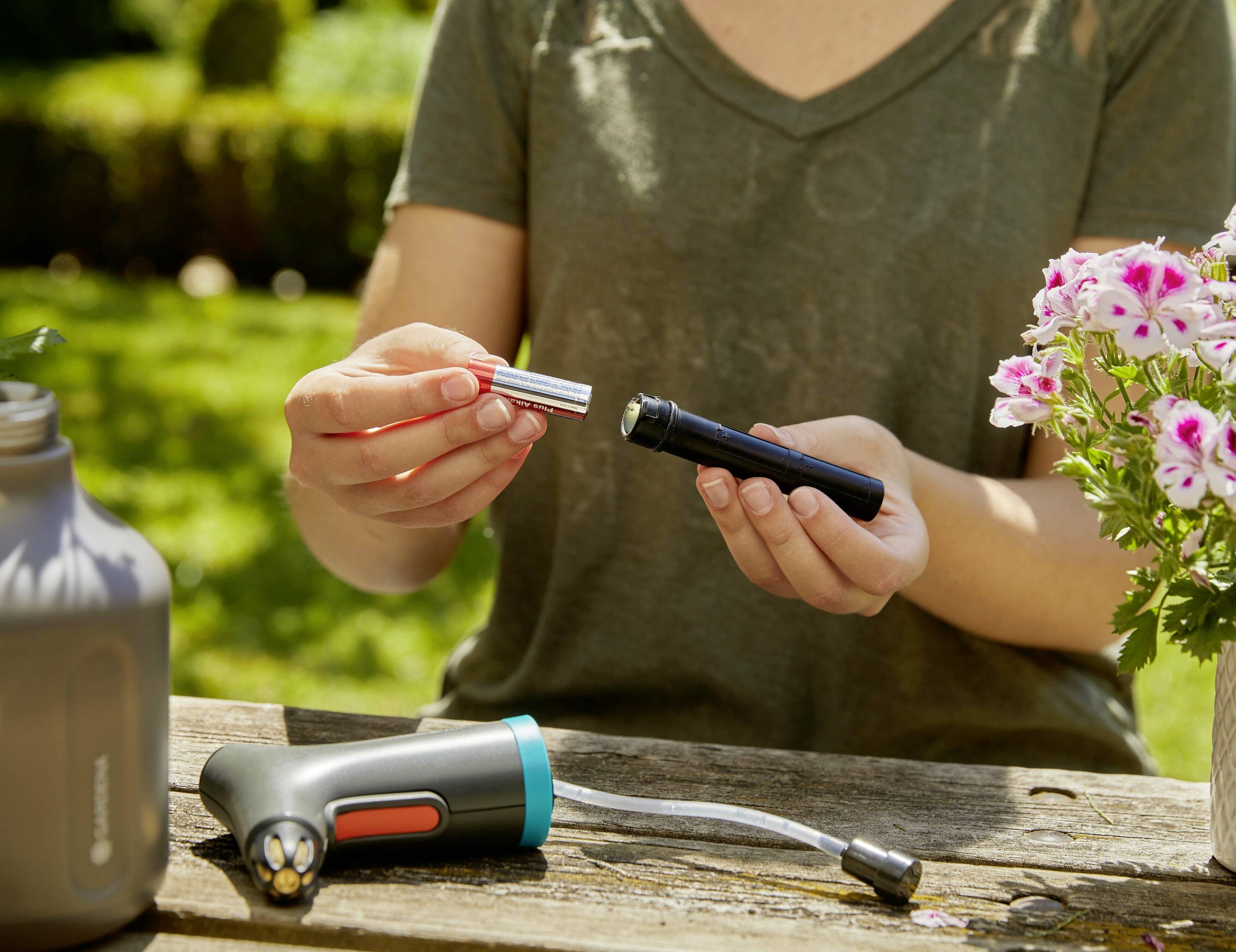 A person is sitting outdoors holding a battery compartment of a portable device. Flowers and another device can be seen on the table.