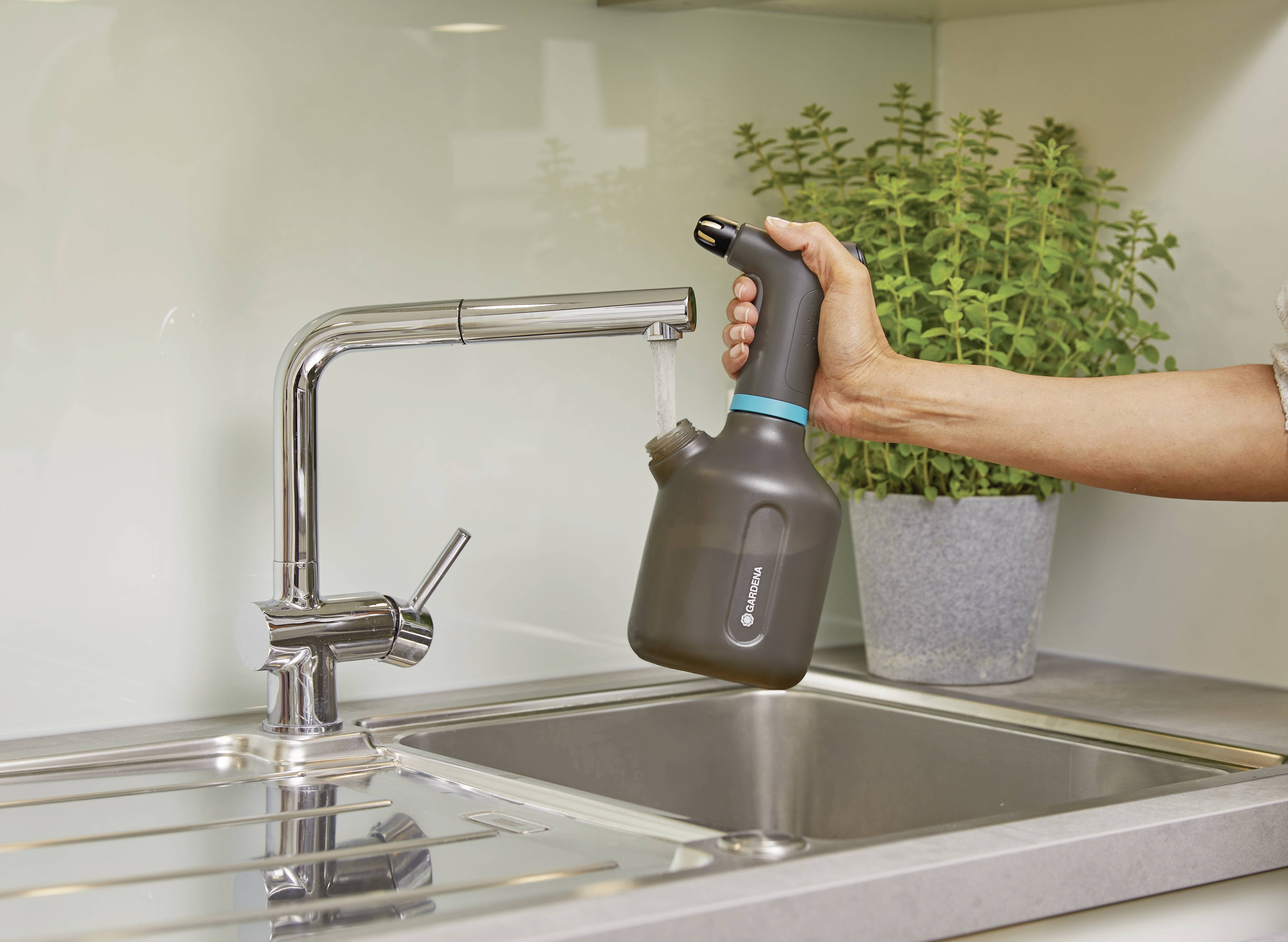 A person is holding a watering can under a tap in a kitchen. A potted plant is in the background.