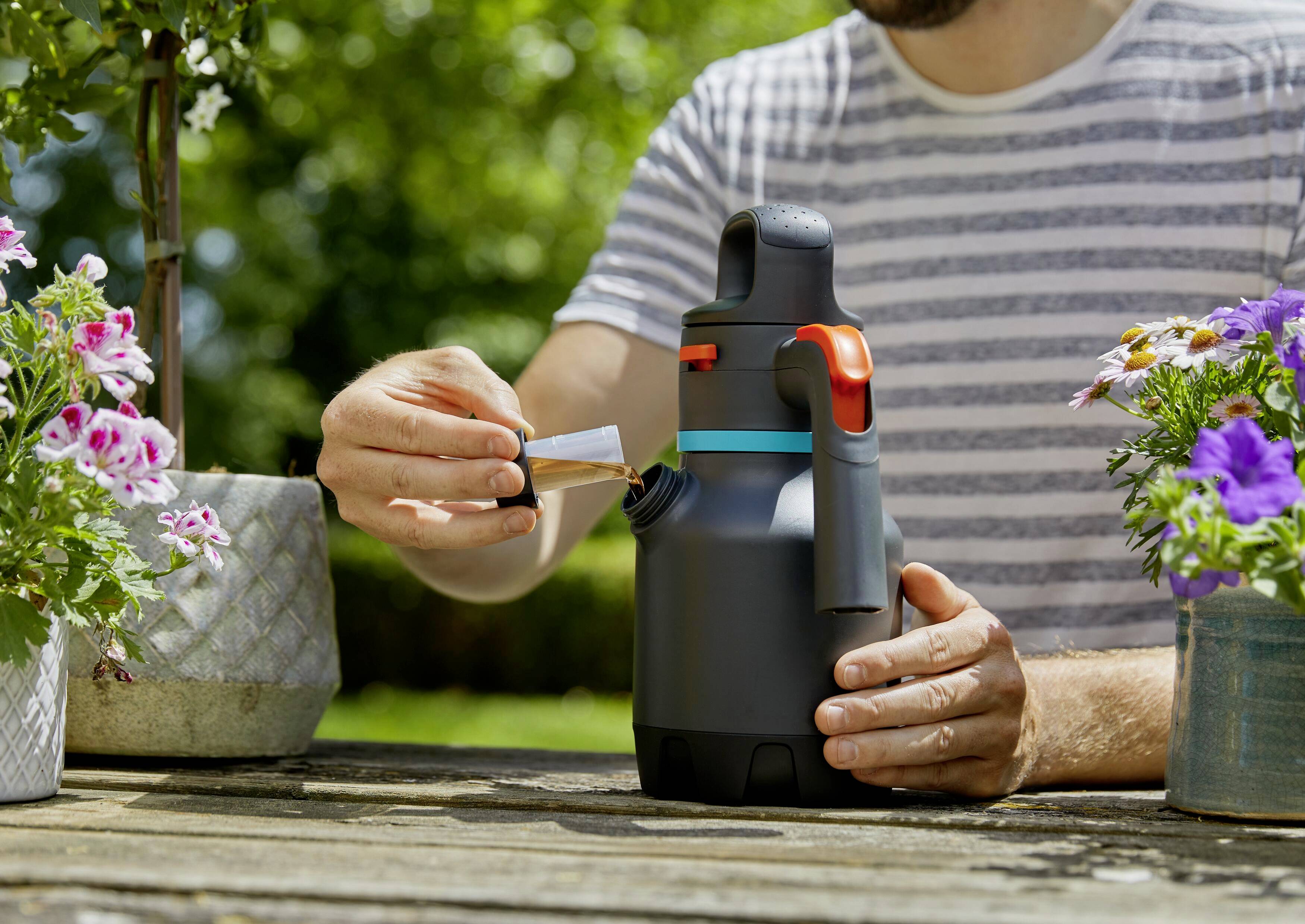 A person is holding a grey watering can with a red handle, pouring fertiliser into it, outside on a wooden table, surrounded by flowers.
