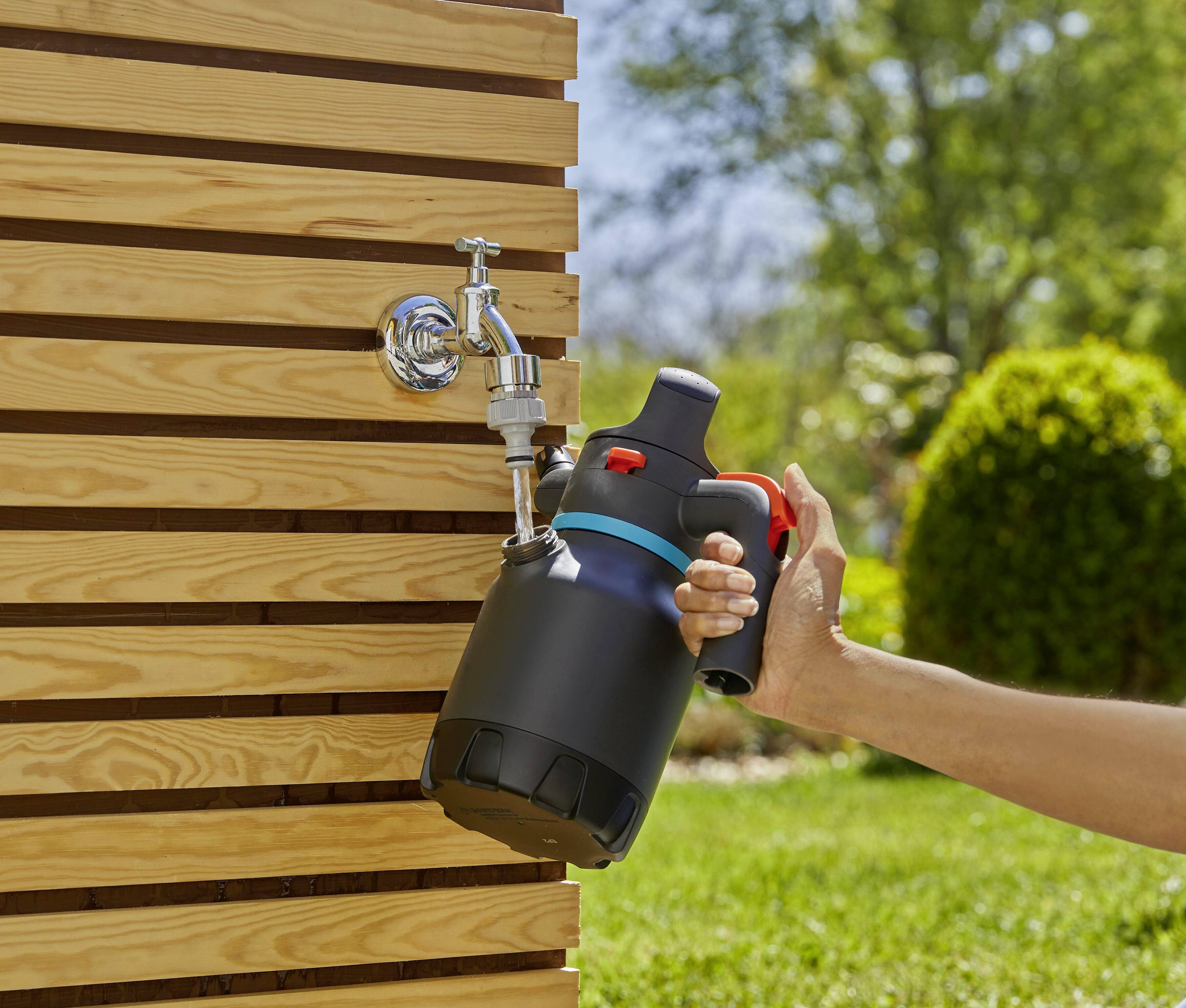 A person is filling a black garden pressure sprayer with water at an outdoor tap, surrounded by a green lawn and sunshine.