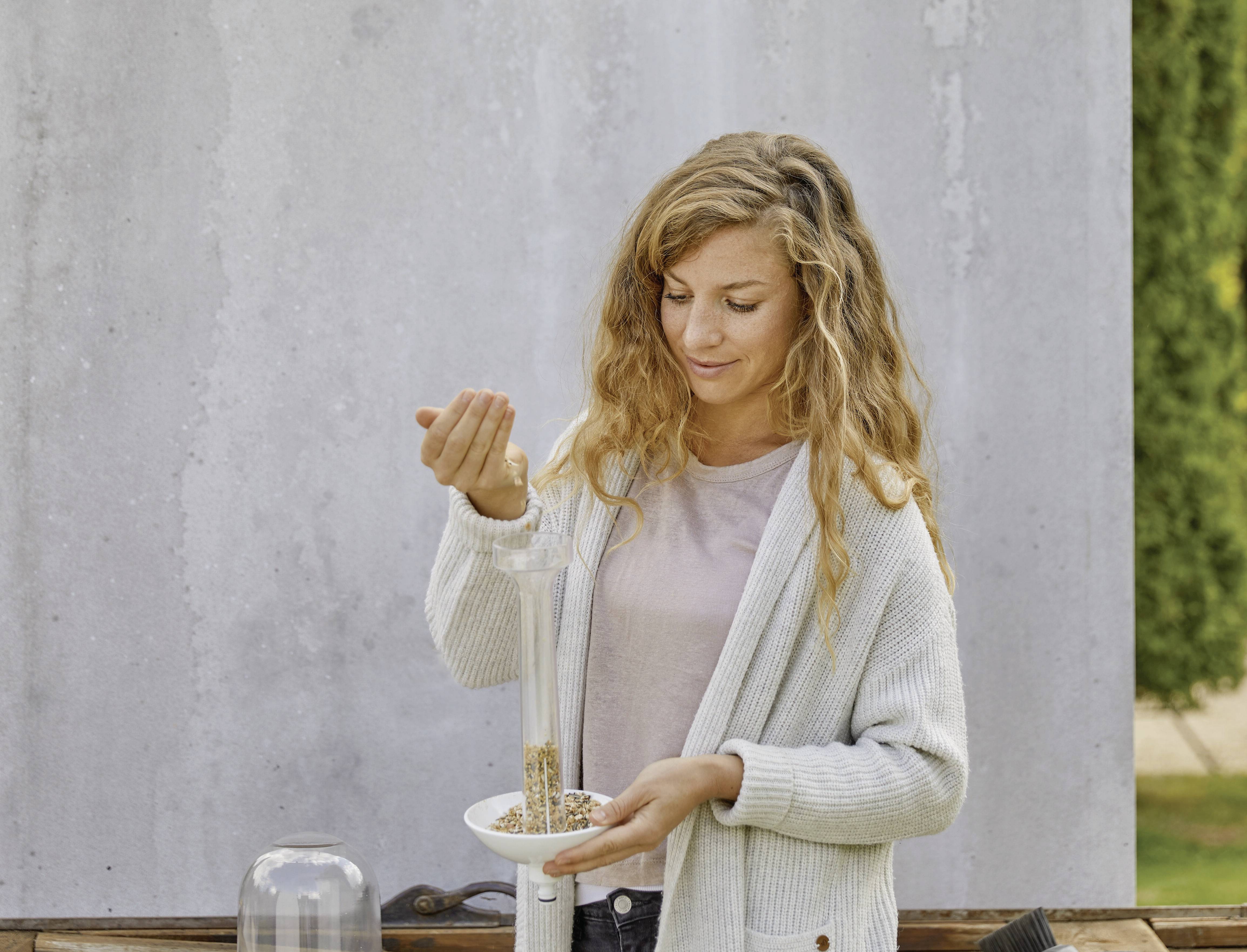 A person is pouring muesli into a glass bowl. They are wearing a light-coloured cardigan and standing in front of a grey wall.