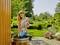 A woman in a garden uses a blue submersible pump in a wooden water barrel, surrounded by flowers and green landscape.