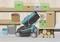 A lawnmower sits in a garage, surrounded by shelves with green and brown boxes, buckets, and stacked logs.