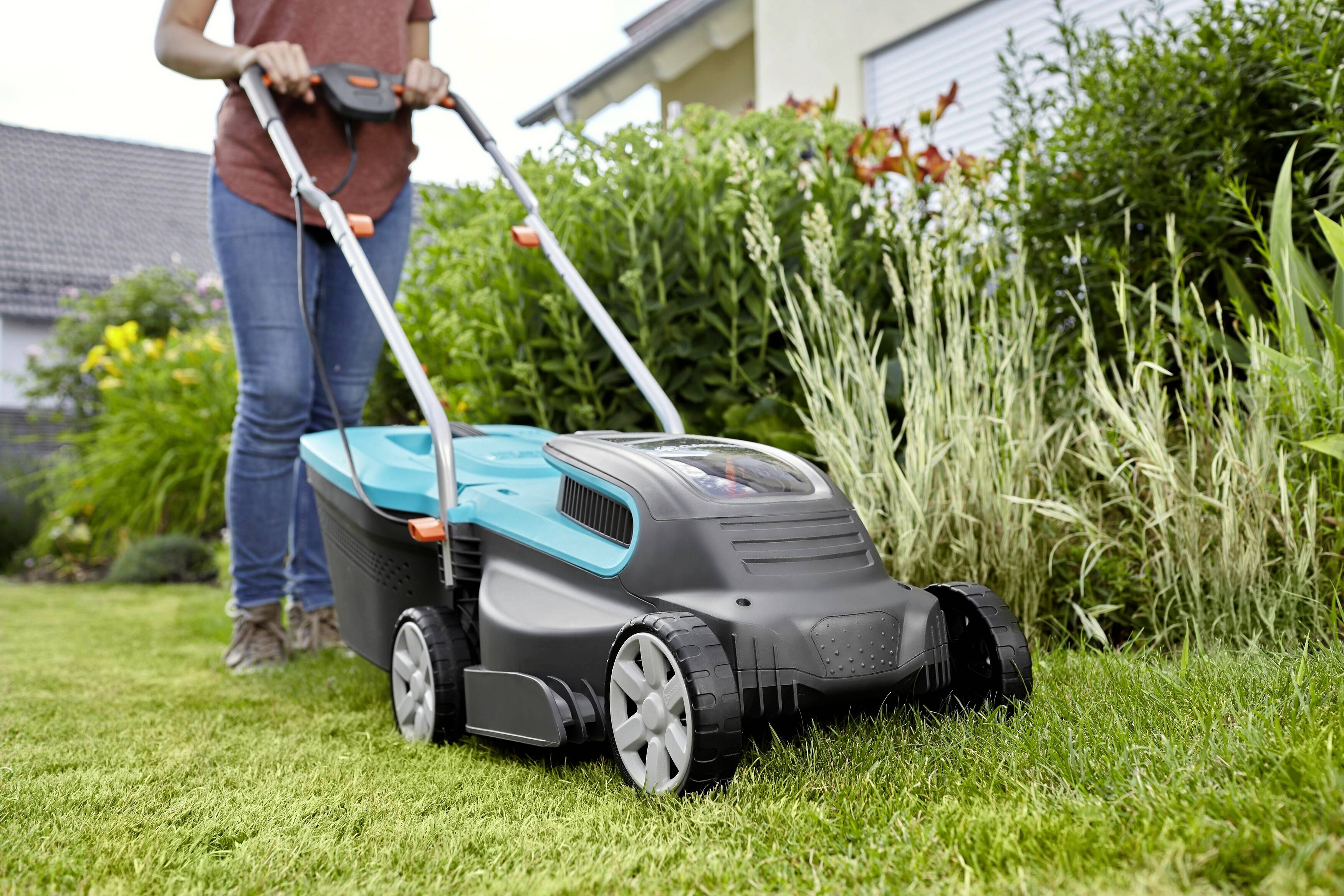 A person is mowing a green lawn with an electric lawnmower. Shrubs and a house are visible in the background.