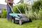 A person is mowing a green lawn with an electric lawnmower. Shrubs and a house are visible in the background.