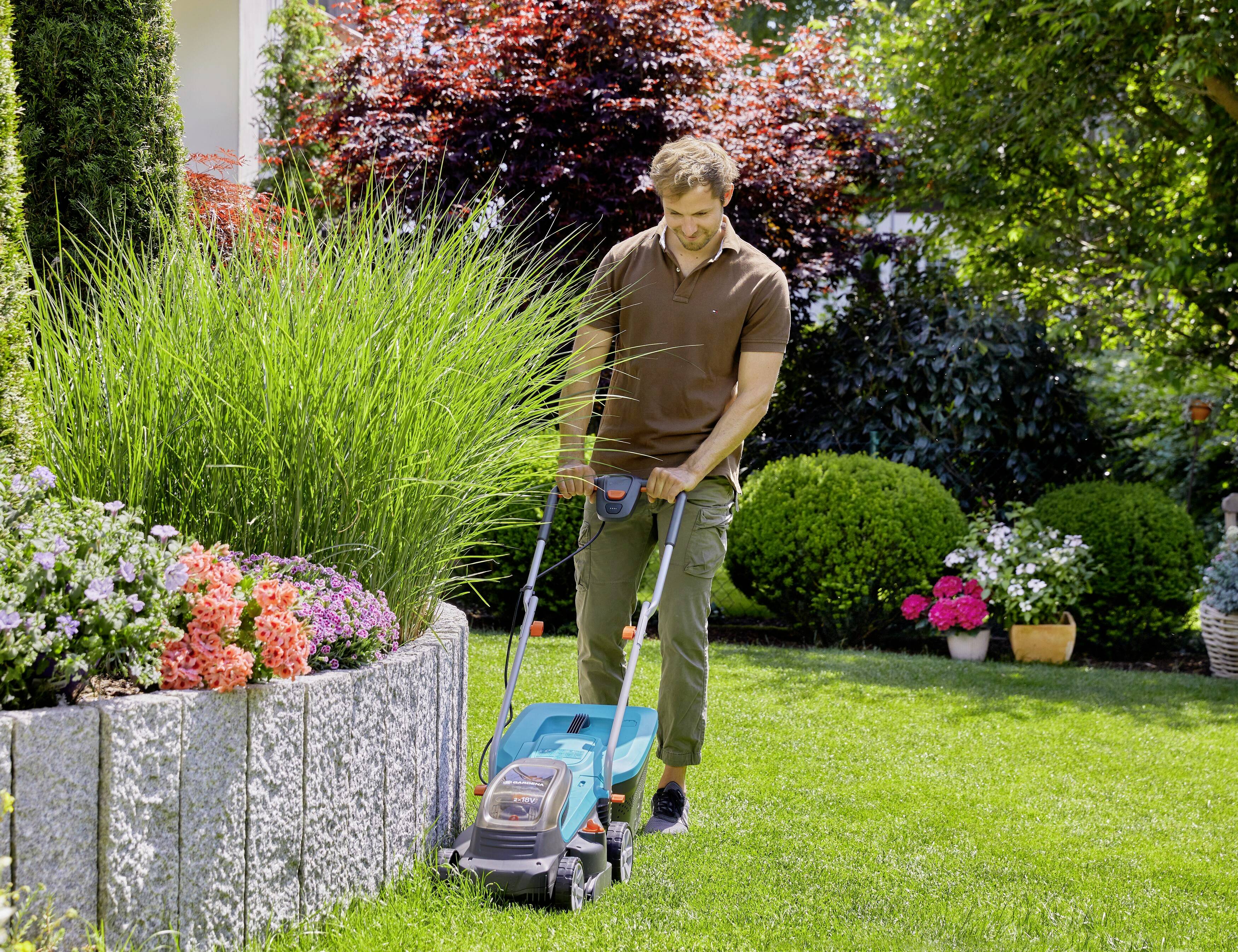 A man is mowing the lawn with an electric lawnmower in a well-maintained garden, surrounded by colourful flowers and shrubs.