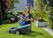 A woman in a garden is assembling a lawnmower. Flowers and shrubs can be seen in the background.