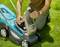 A person is kneeling on a green lawn and holding open the cover of a lawnmower to check or replace the battery.