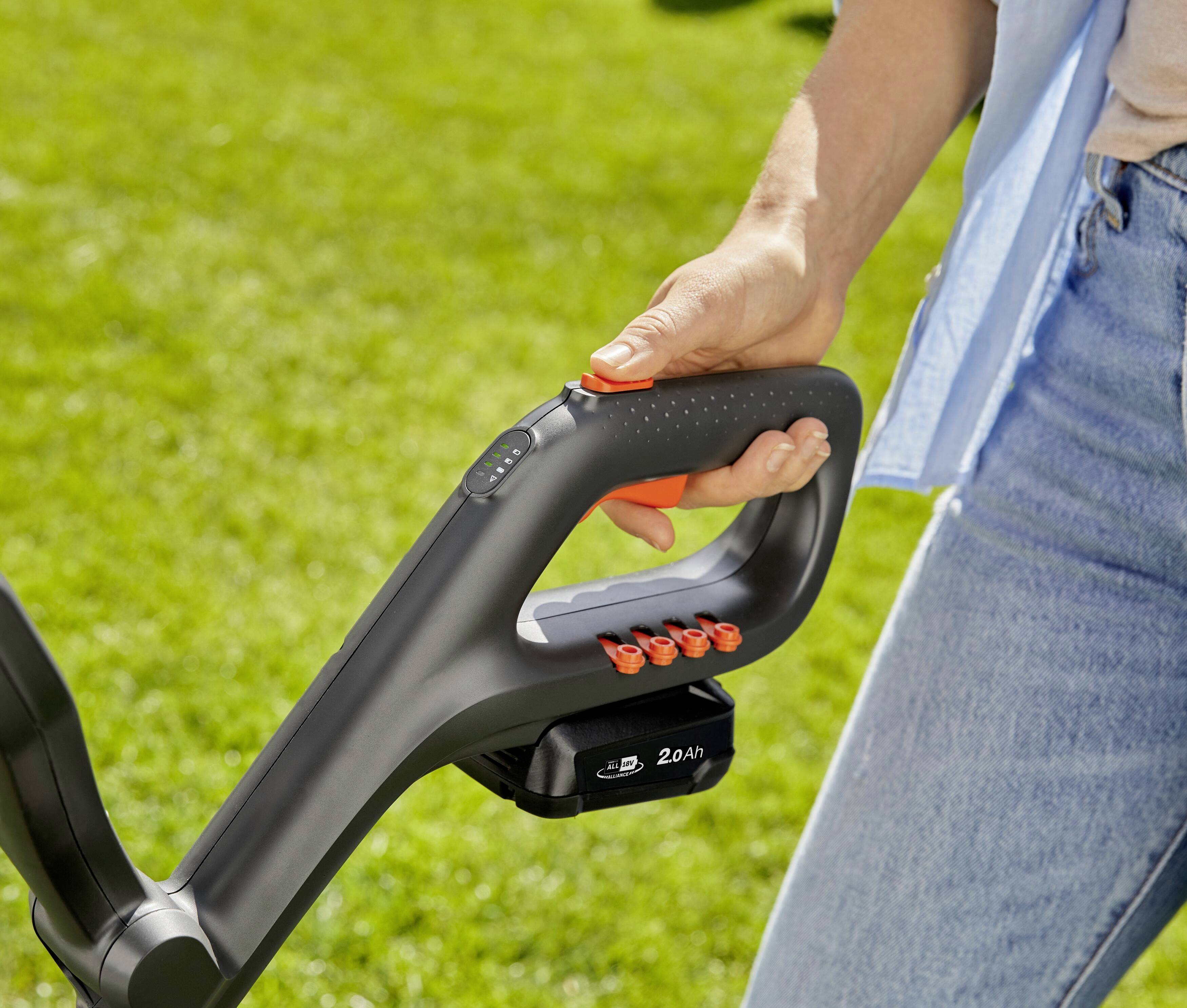 A hand is holding the handle of a cordless electric garden tool on a green lawn. The device has a battery.