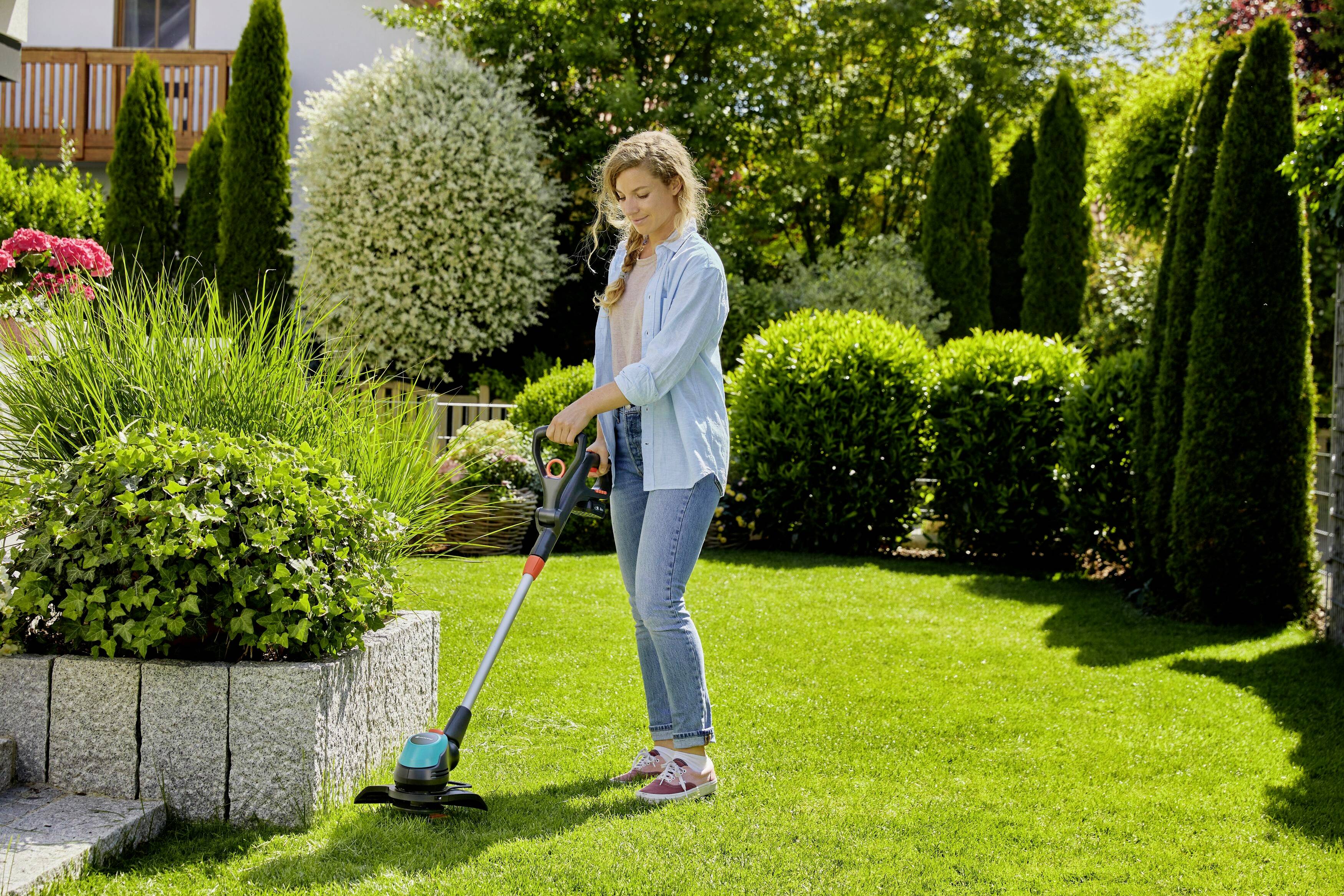 A woman is mowing the lawn with a strimmer in a well-maintained garden. Shrubs and trees can be seen in the background.