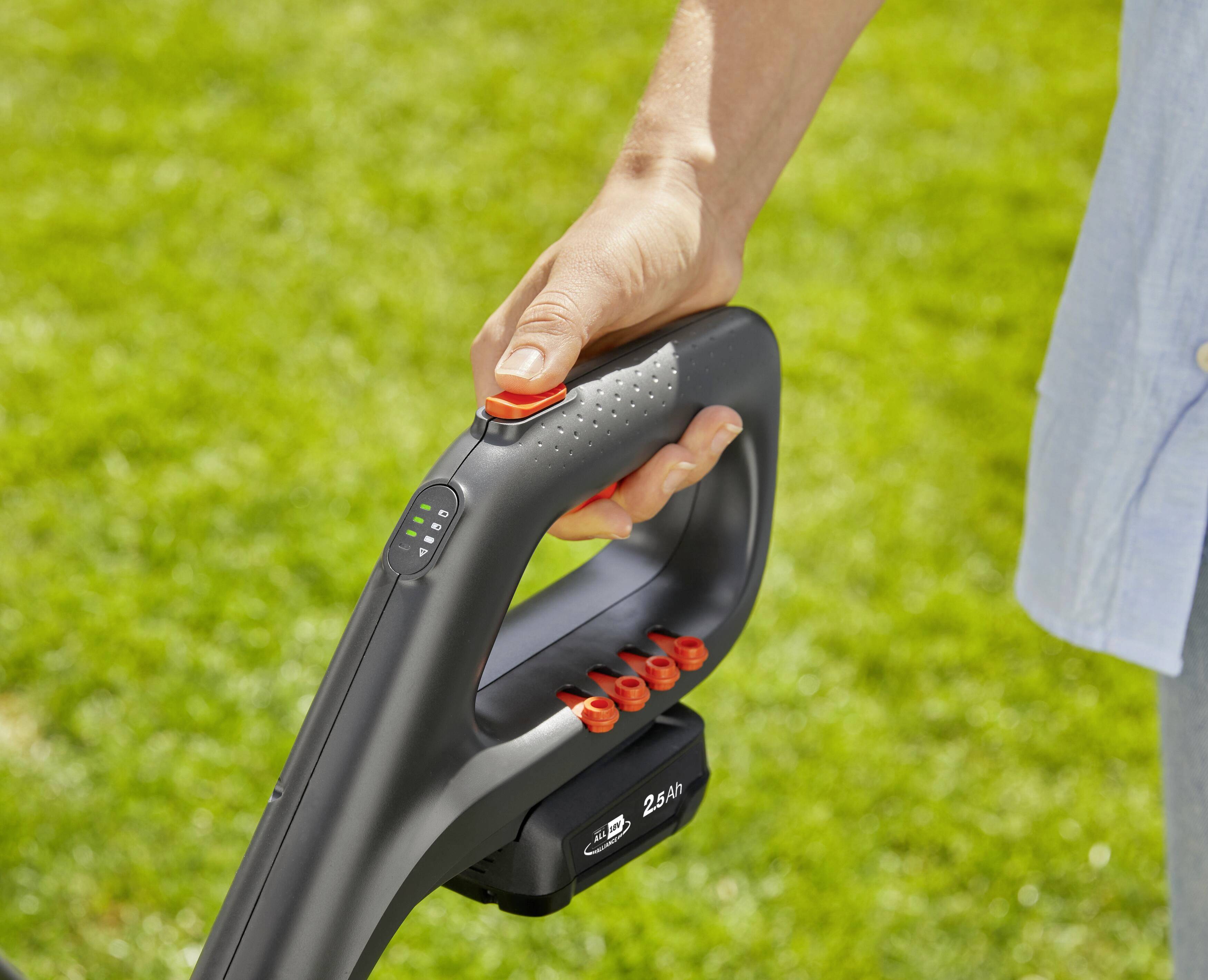 A person is holding the handle of a lawnmower, pressing a red button. The background shows green grass.